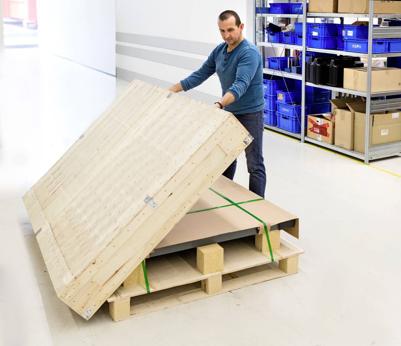 Un homme soulève prudemment une grande caisse en bois d'une palette dans une zone d'entreposage, avec des cartons et des sangles en dessous.