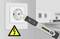 Dans une pièce, une prise électrique est testée pour la présence de tension à l'aide d'un testeur de tension. Un panneau d'avertissement signale un danger électrique.
