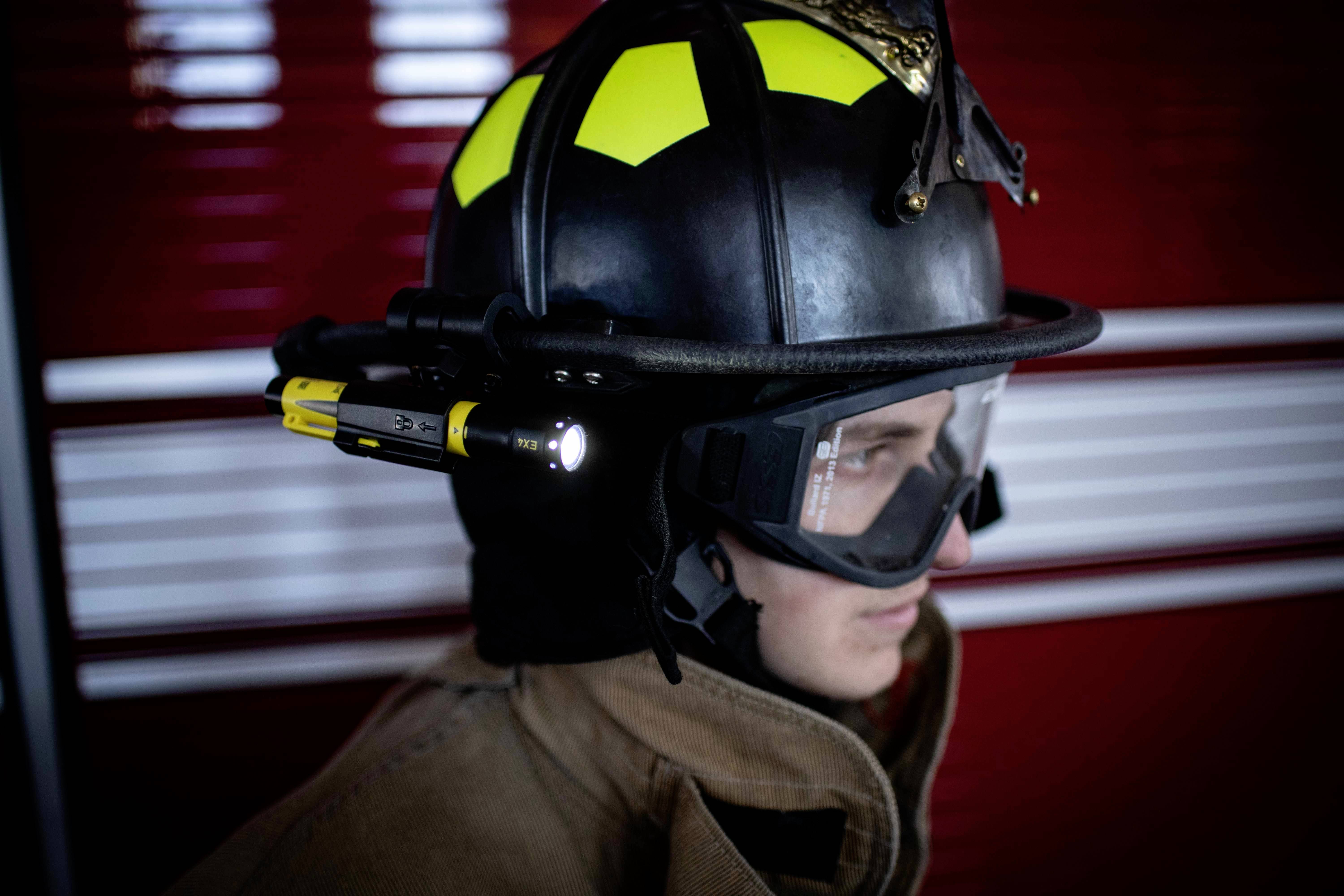 Un pompier de profil porte un casque avec lampe intégrée, des lunettes de protection et une tenue de protection devant un fond rouge.