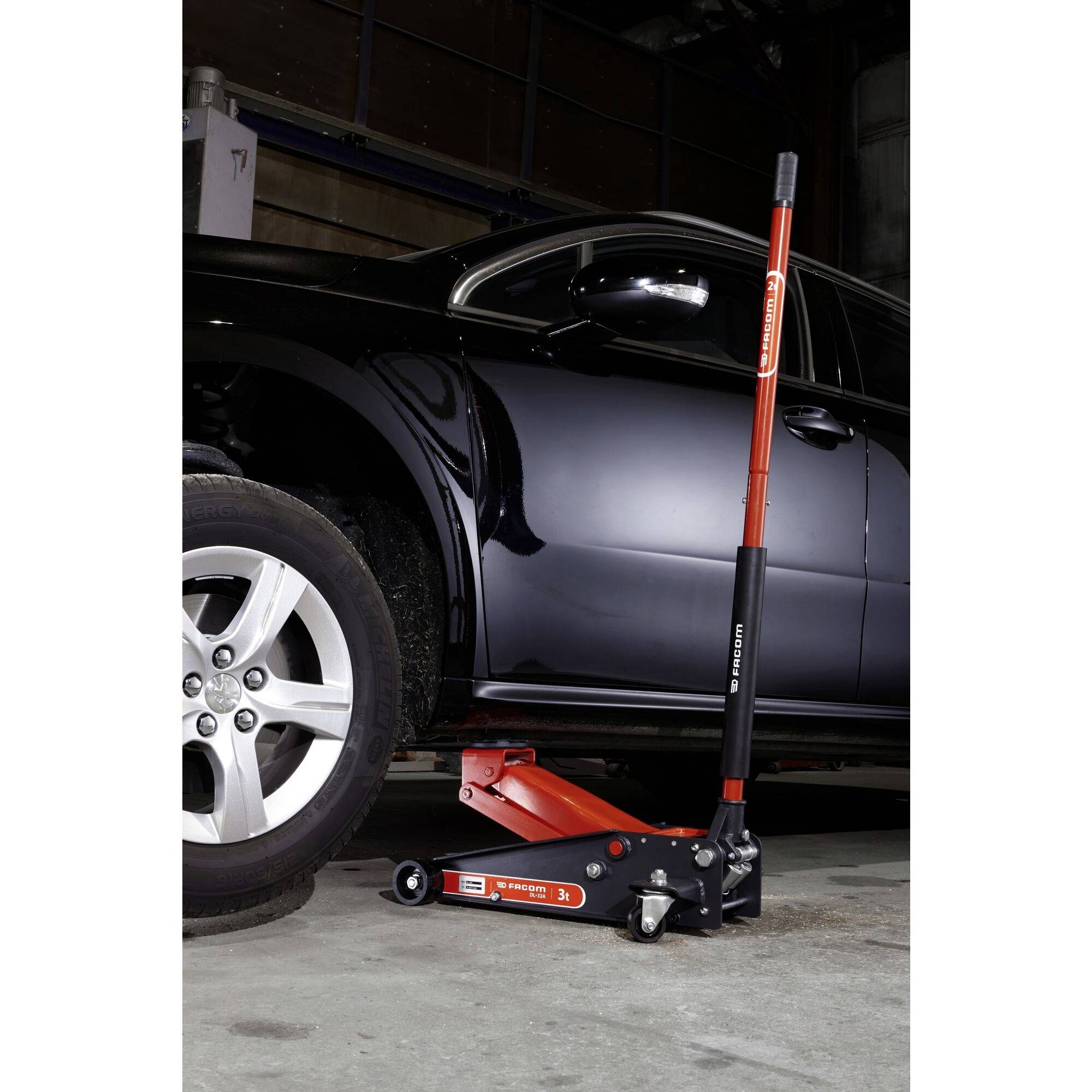 Une voiture noire est soulevée dans un garage à l'aide d'un cric hydraulique. Le cric est rouge et noir.