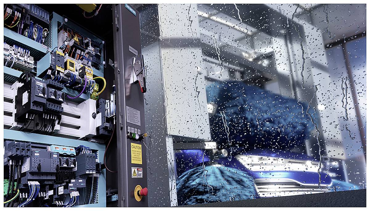 Un système industriel aux circuits ouverts est au premier plan. Derrière lui, une fenêtre avec des gouttes de pluie donnant sur des machines.