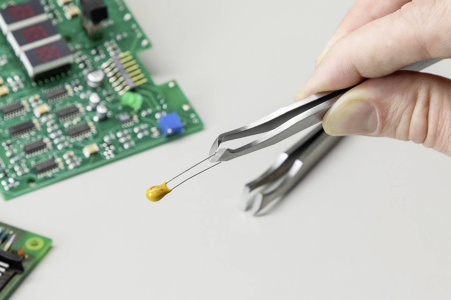 Une main tient avec une pince un petit point de colle au-dessus d'une carte de circuit imprimé équipée de composants électroniques.