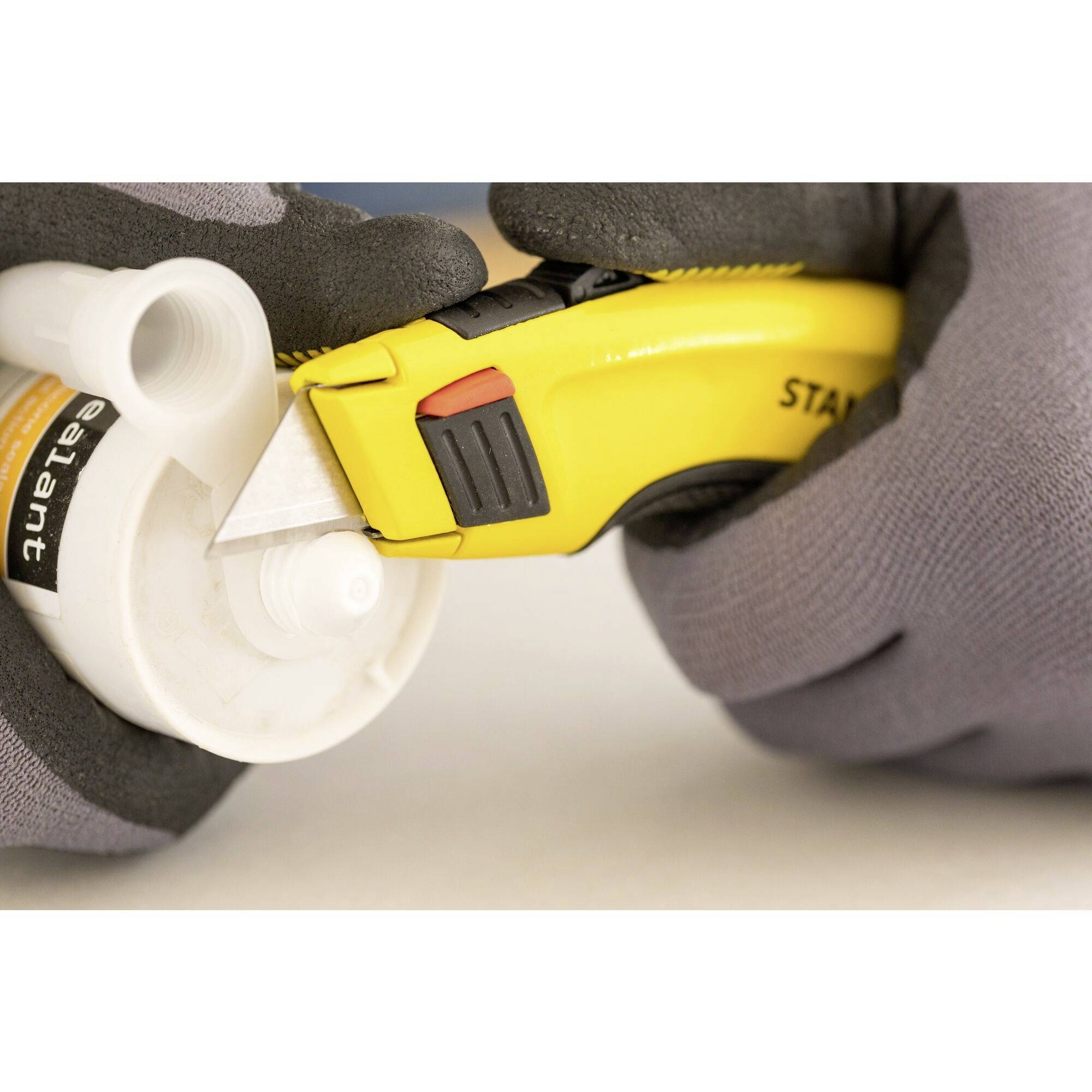 Une personne portant des gants gris coupe la pointe d'un récipient de colle avec un cutter jaune.