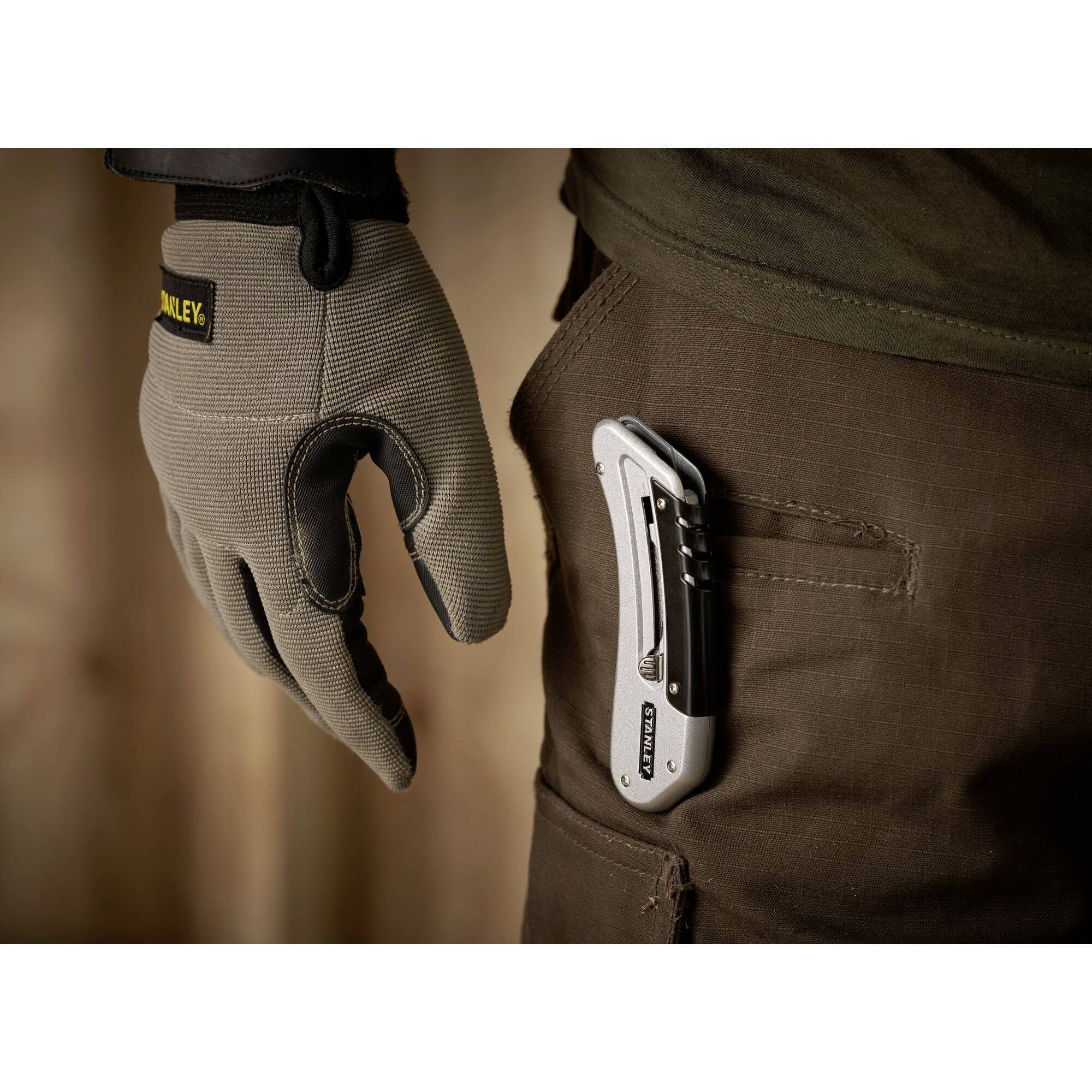 Une main gantée de gants de travail tient un couteau de poche fermé. Un autre couteau de poche est visible dans la poche arrière du pantalon.
