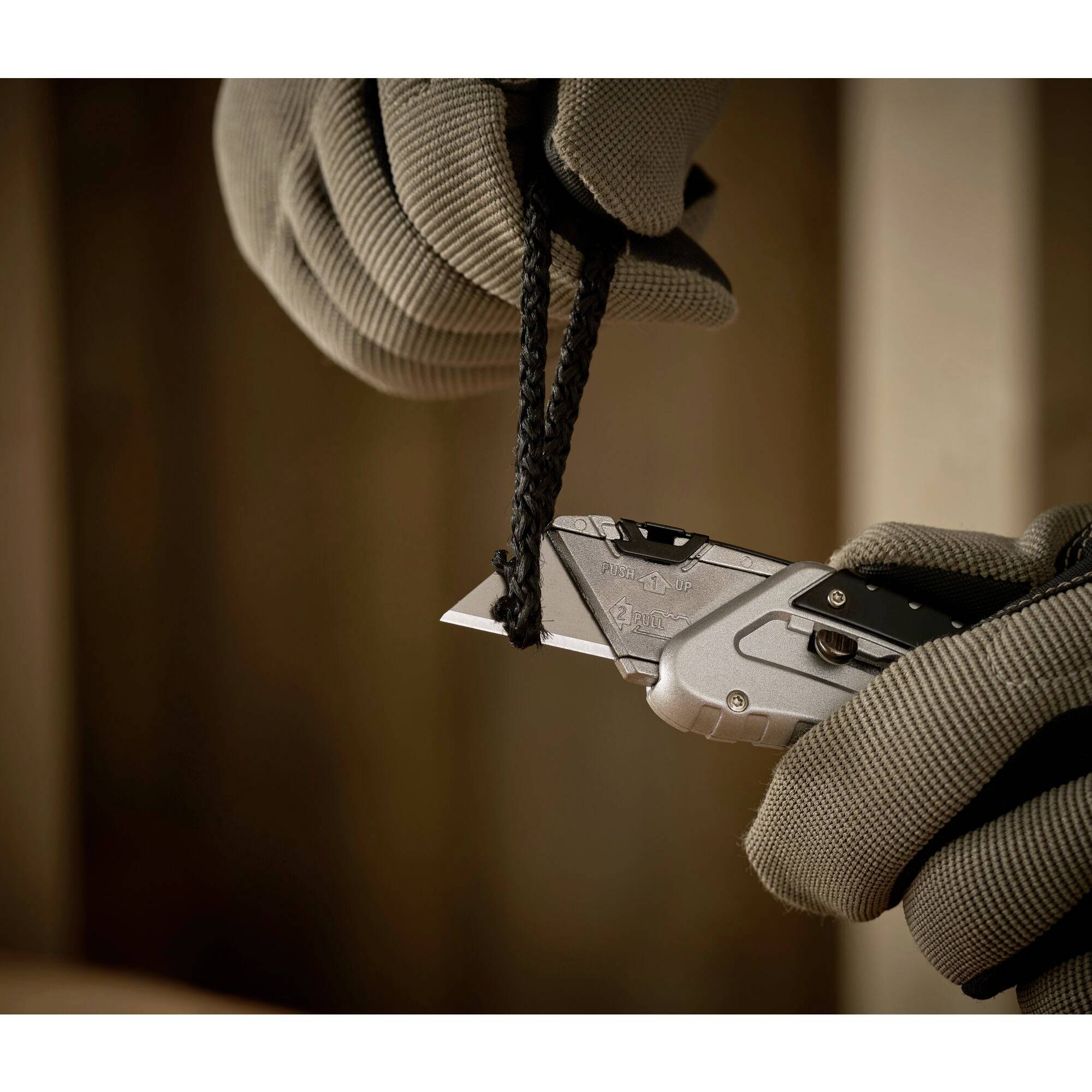 Une personne portant des gants gris coupe une corde noire avec un couteau pliant argenté. L'arrière-plan est flou, avec le focus sur le couteau et la corde.