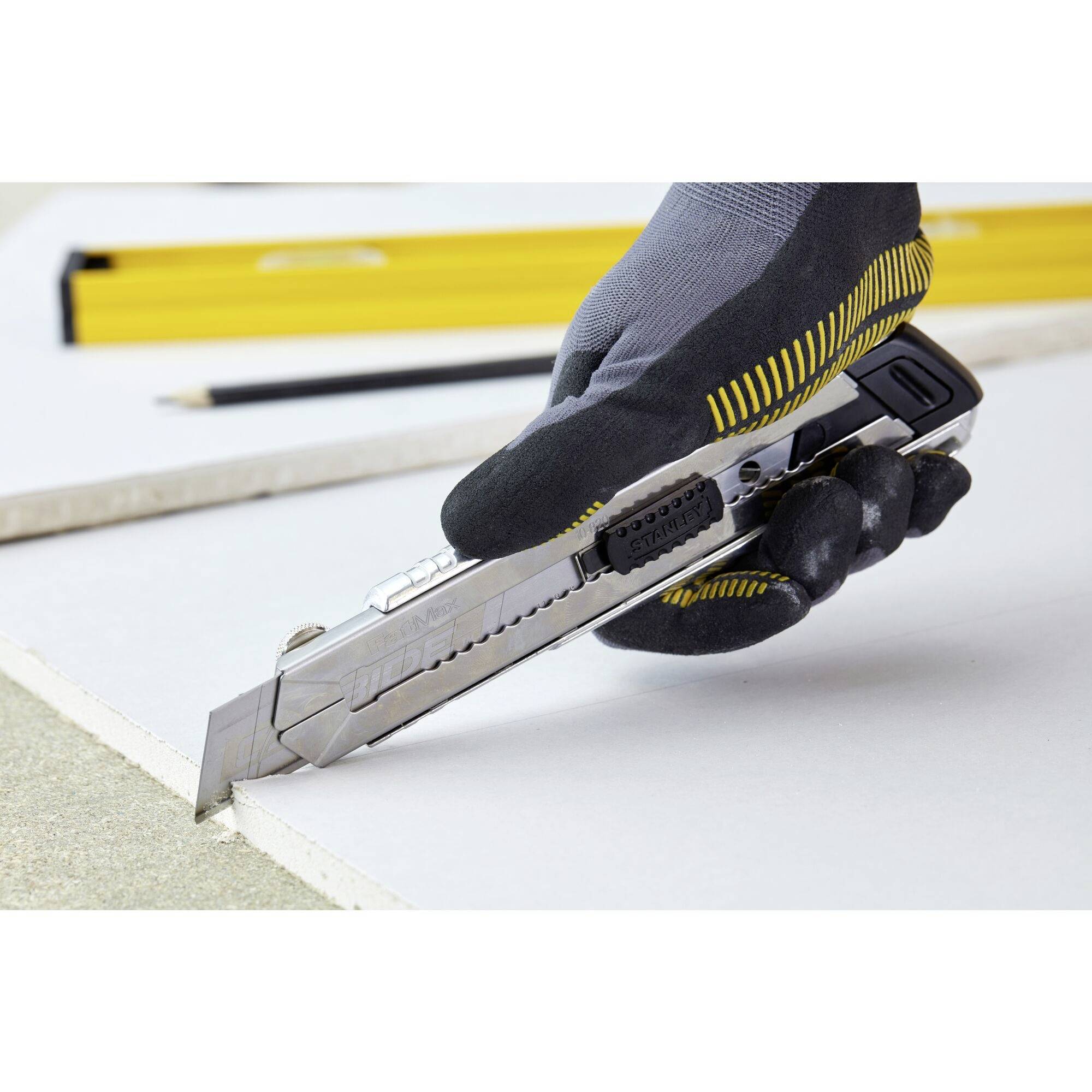 Une main gantée tient un cutter et coupe précisément une surface de plaque blanche. Une règle jaune est posée à côté.