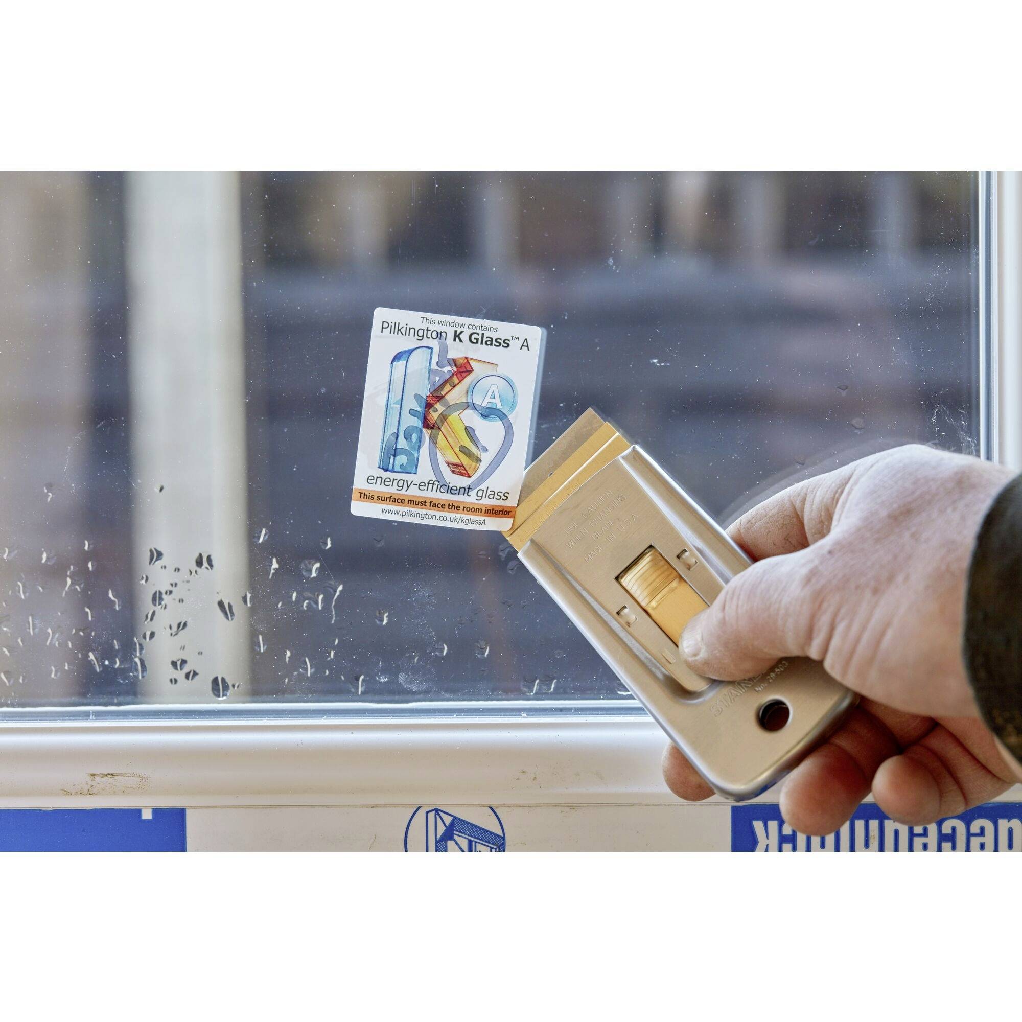 Une personne retire une étiquette d'une fenêtre. Sur l'étiquette, il est écrit 'Pilkington K Glass™ A'. La fenêtre indique du 'verre à faible consommation d'énergie'.
