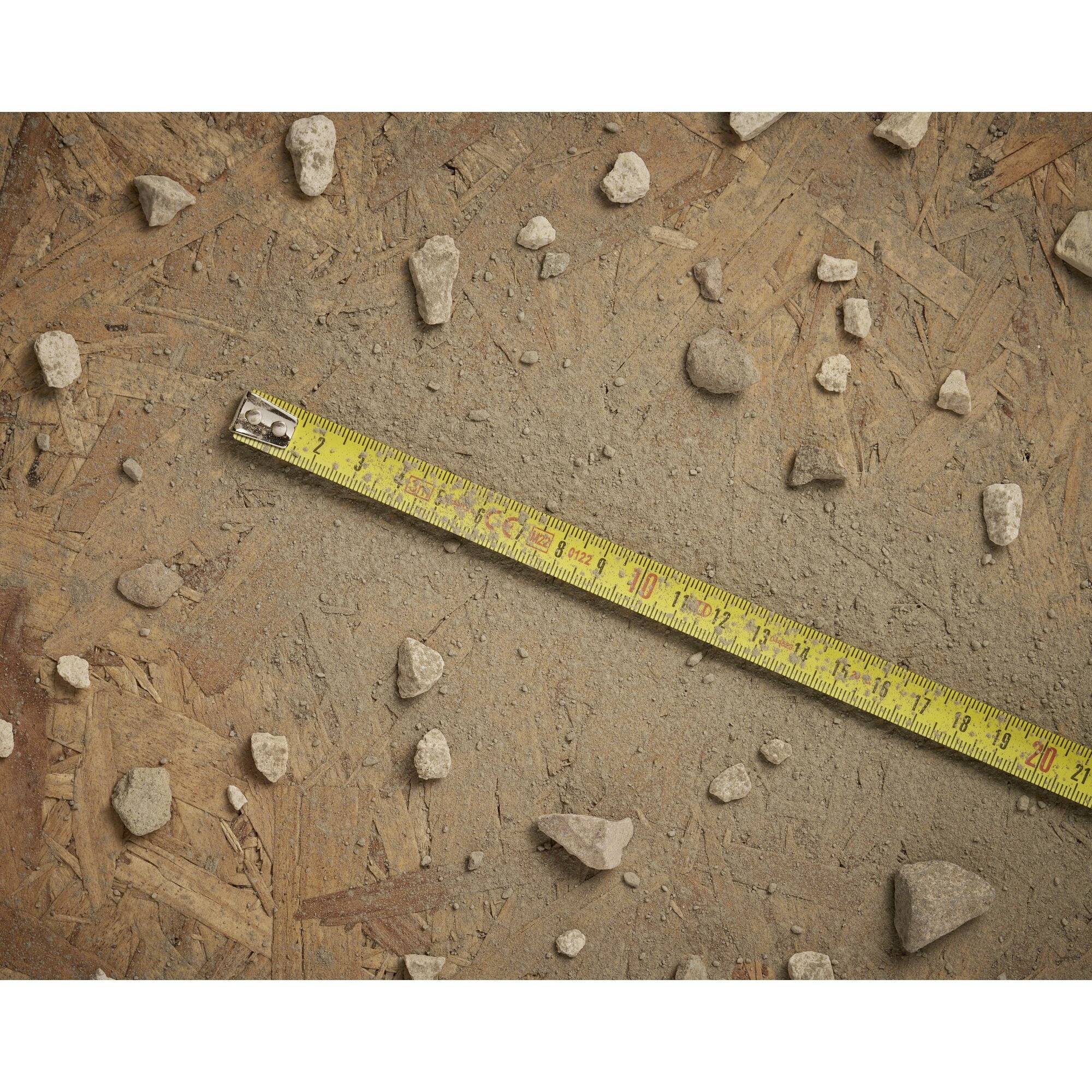 Un mètre ruban jaune est posé diagonalement sur un sol de matériau ressemblant à du panneau de particules, entouré de petites pierres dispersées.