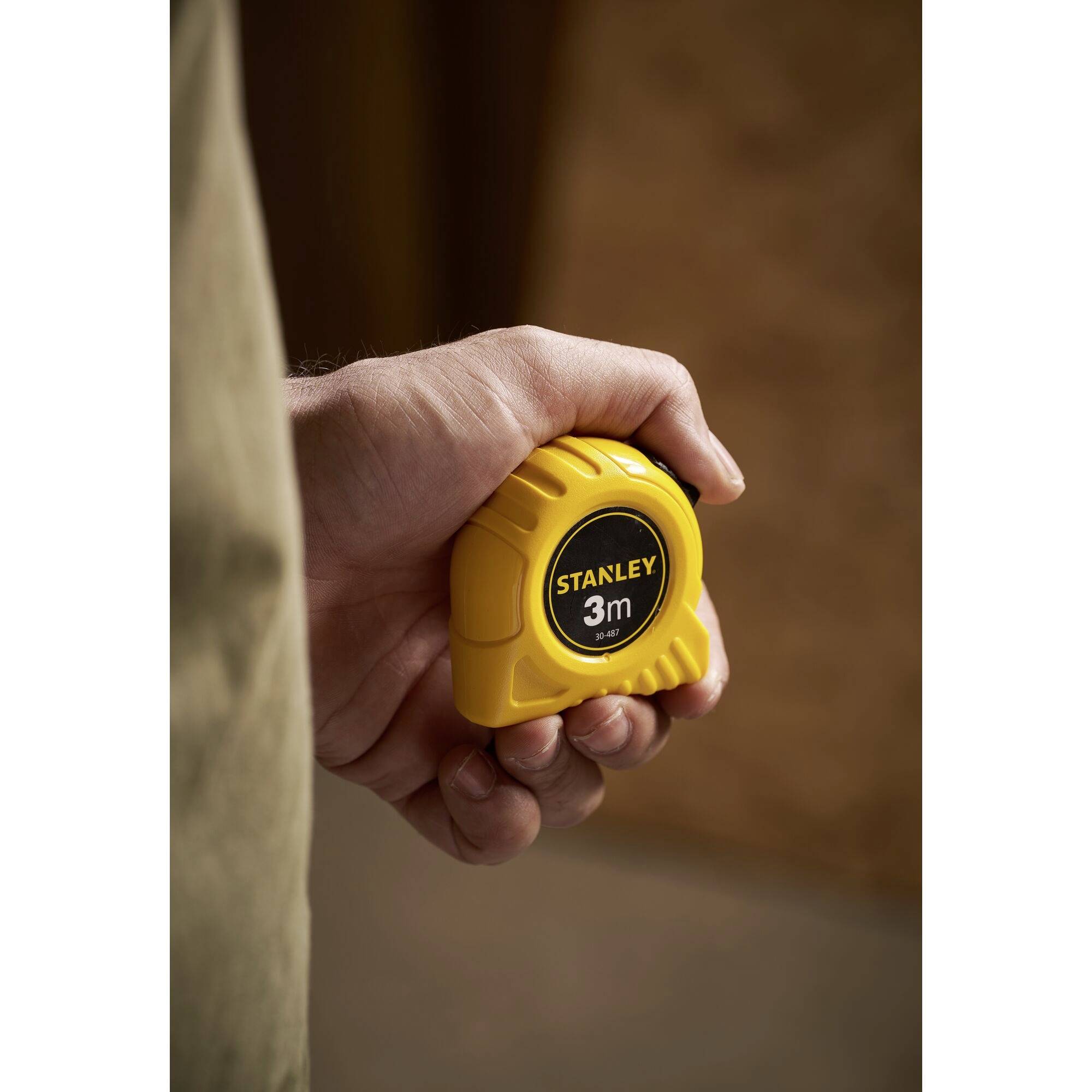 Une main tient un mètre ruban jaune de la marque Stanley avec l'inscription '3m'.