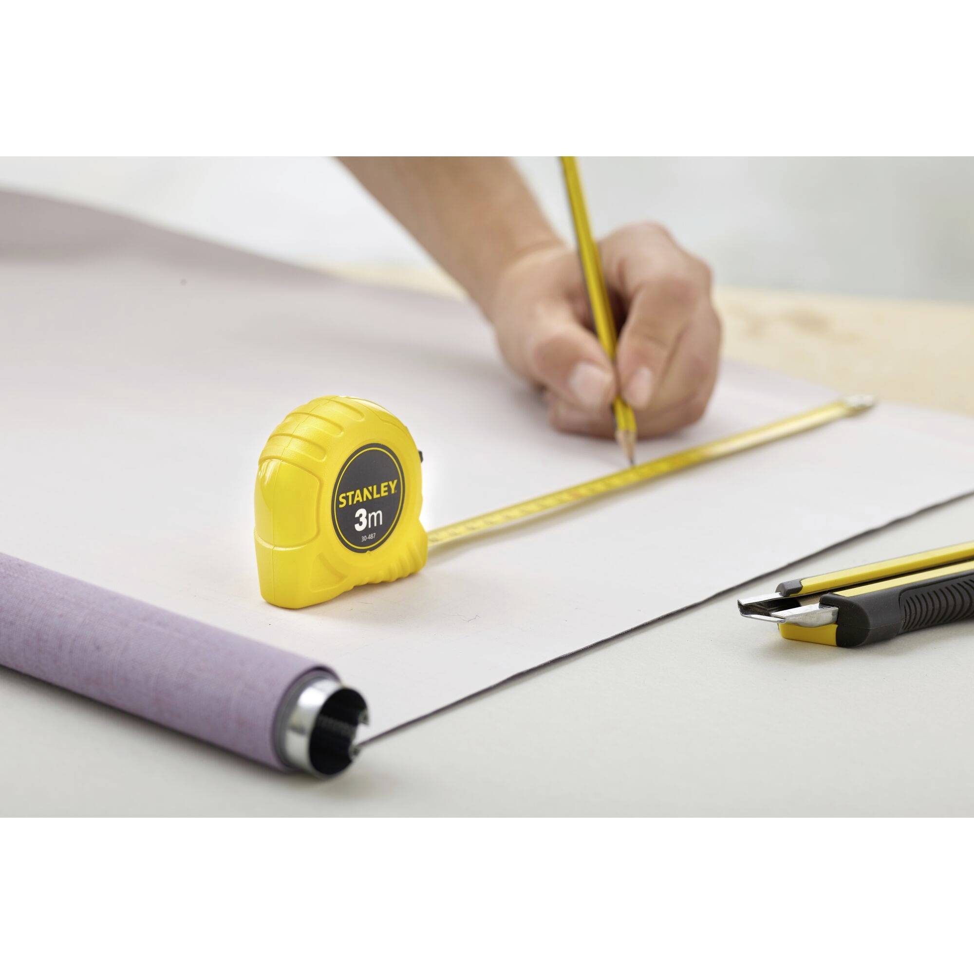 Une main traçant une ligne avec un crayon jaune sur du papier blanc, à côté d'un mètre ruban jaune de 3m posé sur une table.