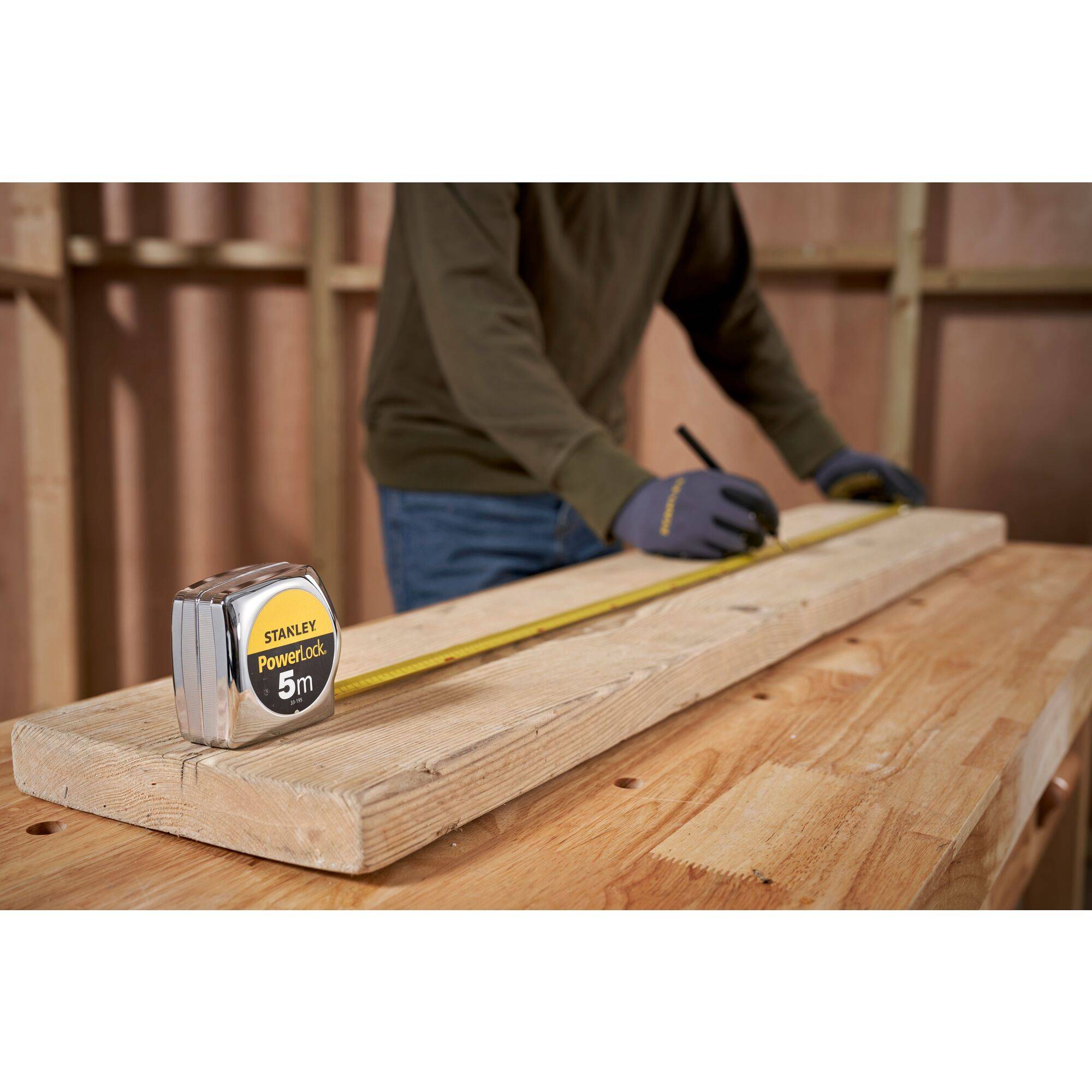 Une personne recherche une planche en bois avec un mètre ruban sur un établi. Un cadre en bois en arrière-plan suggère un environnement de construction.