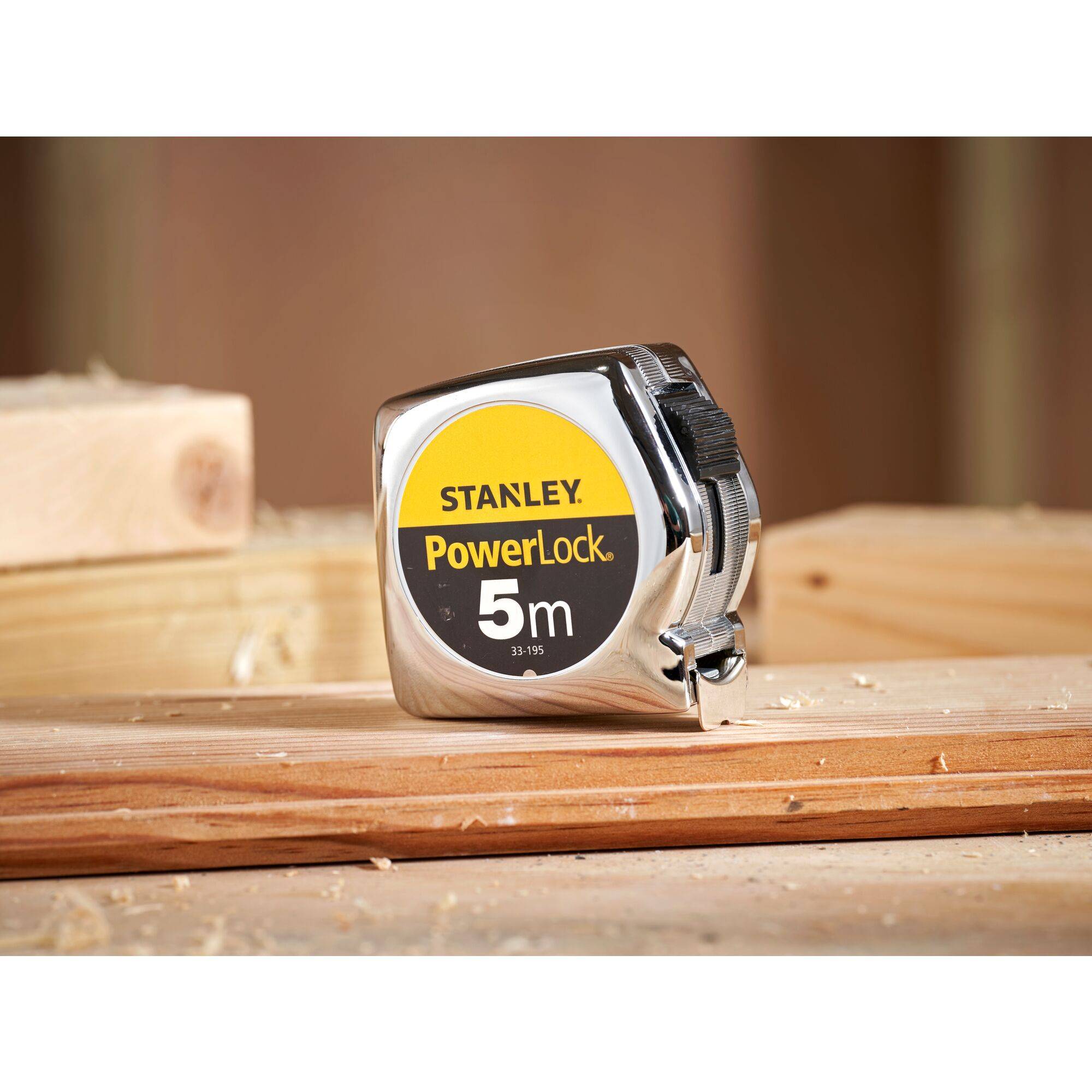 Un ruban à mesurer en métal avec l'étiquette jaune 'Stanley PowerLock 5m' est posé sur une poutre en bois. À l'arrière-plan, on aperçoit d'autres morceaux de bois.