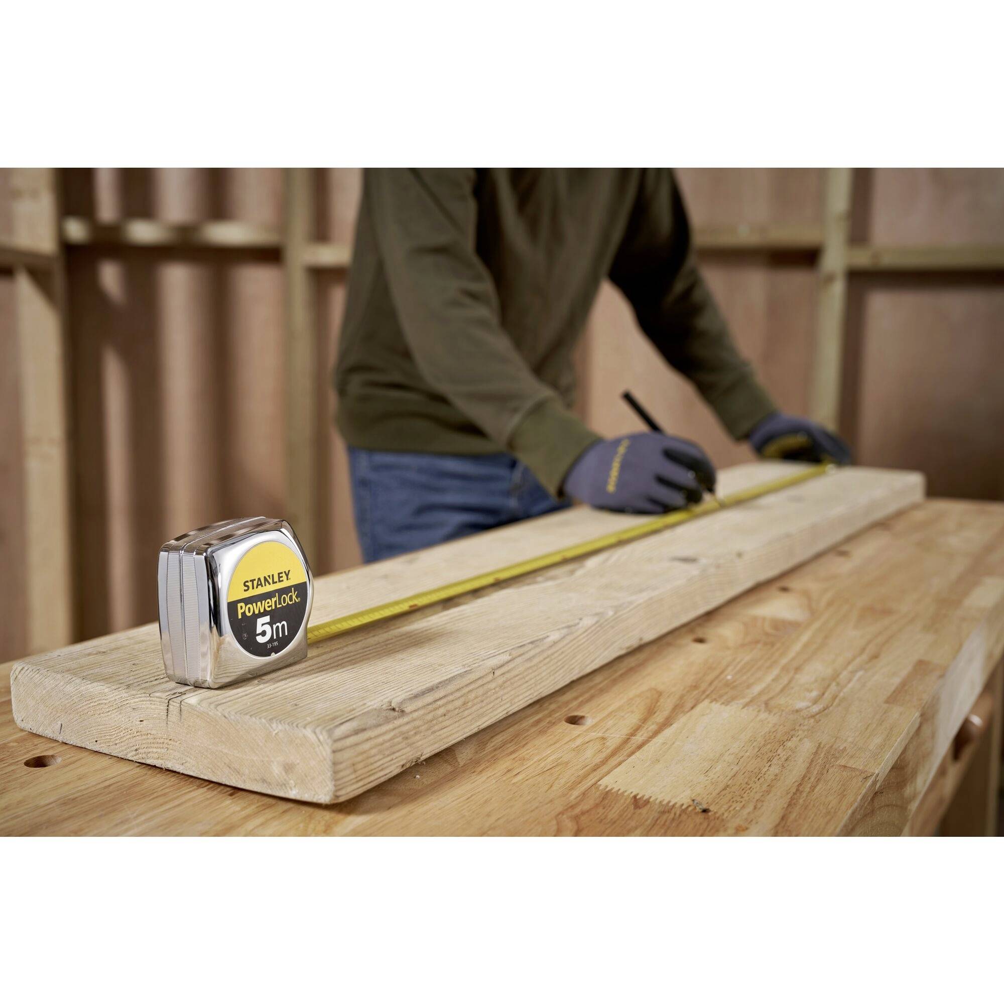 Une personne mesure une planche en bois à l'aide d'un mètre ruban. La personne porte des gants et marque un repère sur la planche.