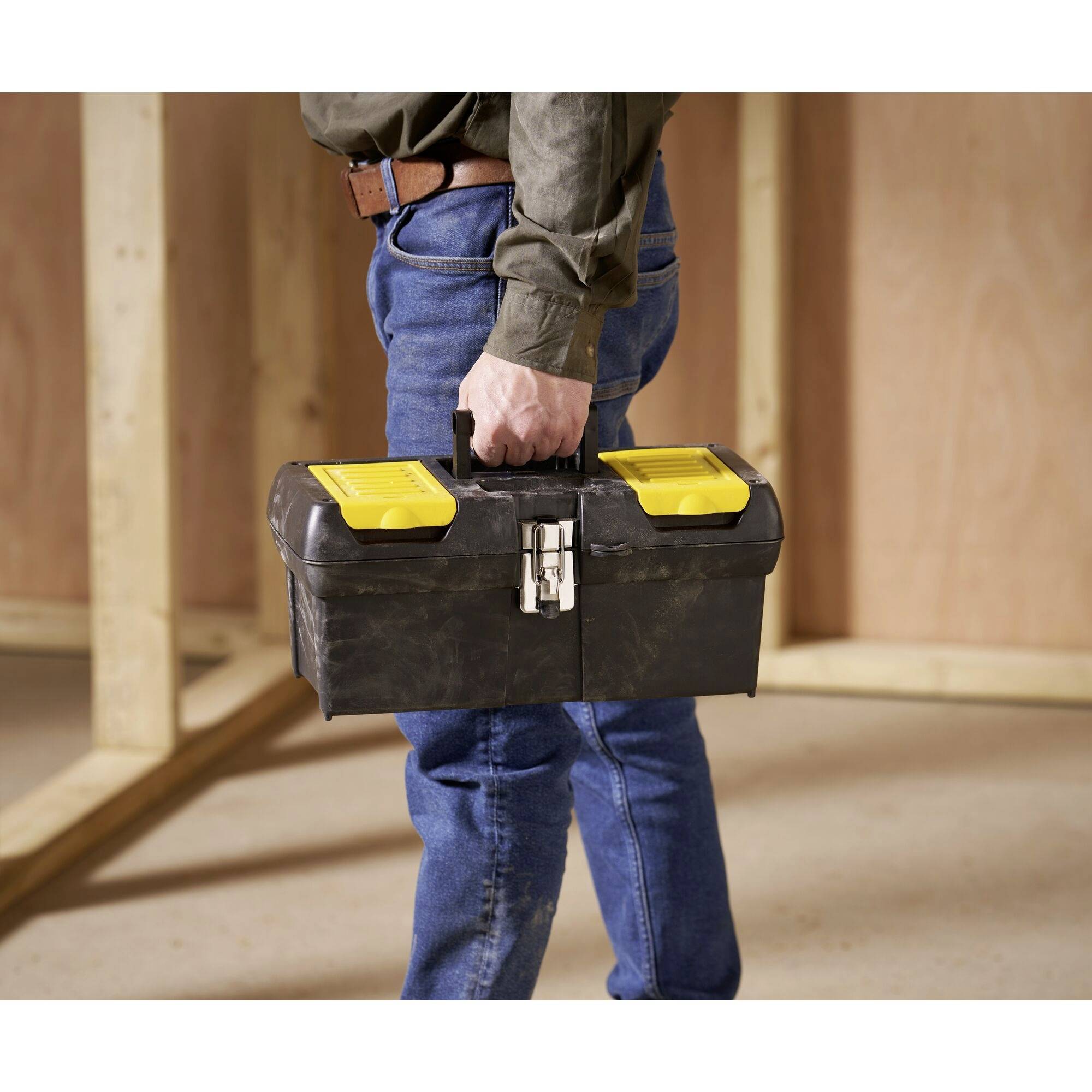Une personne portant un jean et une chemise transporte une boîte à outils noire avec des fermoirs jaunes. L'arrière-plan montre des murs en bois non finis.