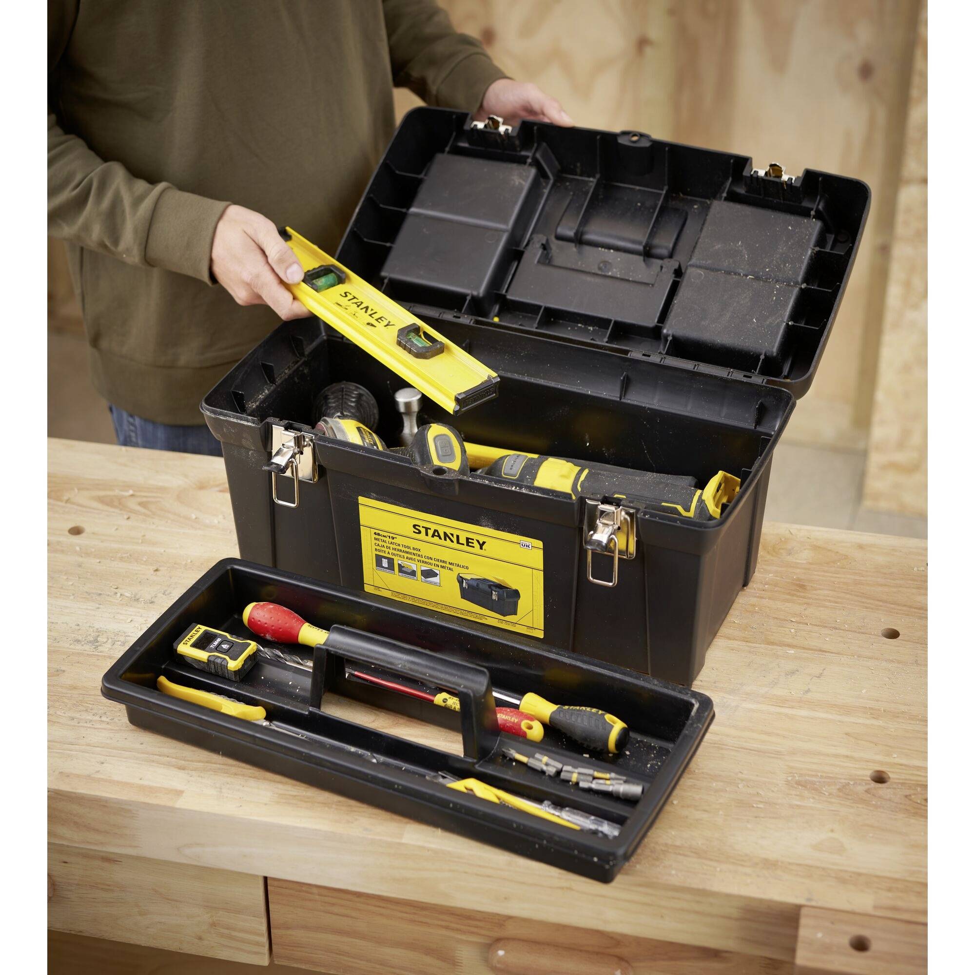 Une personne ouvre une boîte à outils sur un établi. Divers outils, dont des niveaux à bulle et des tournevis, sont visibles.