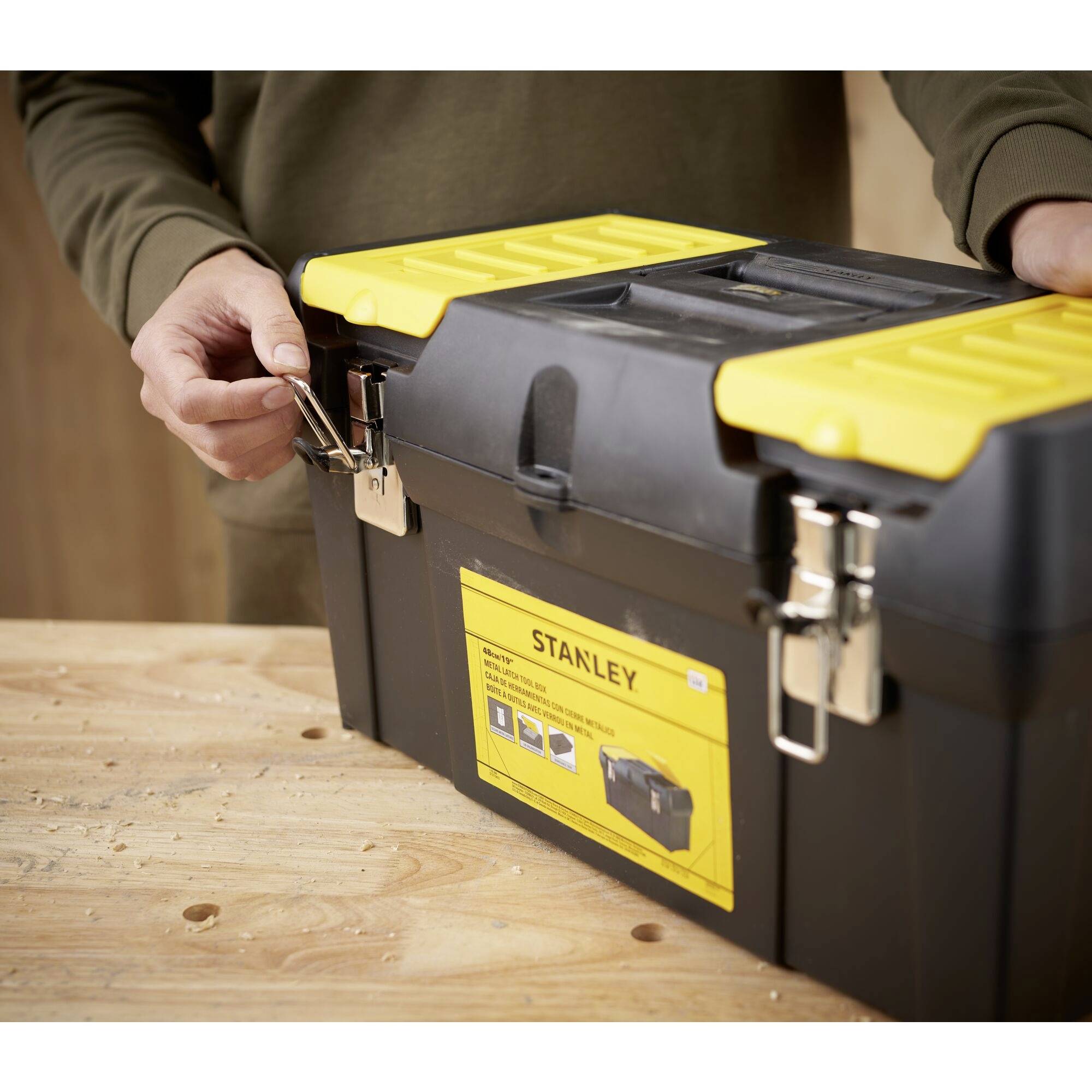 Une personne ouvre une boîte à outils noire avec un couvercle jaune sur une table. Une étiquette portant l'inscription 'Stanley' est visible.