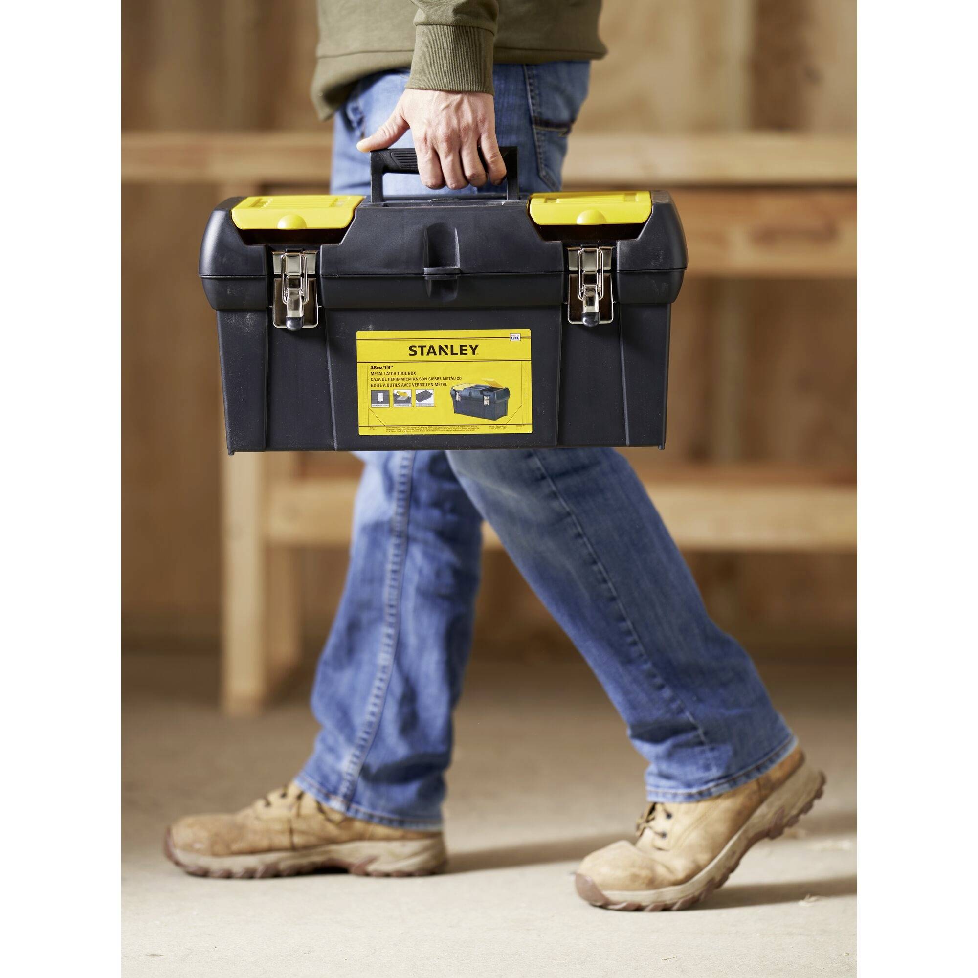 Un homme porte une boîte à outils noire avec une étiquette jaune sur laquelle est écrit 'Stanley'. En arrière-plan, des étagères en bois.