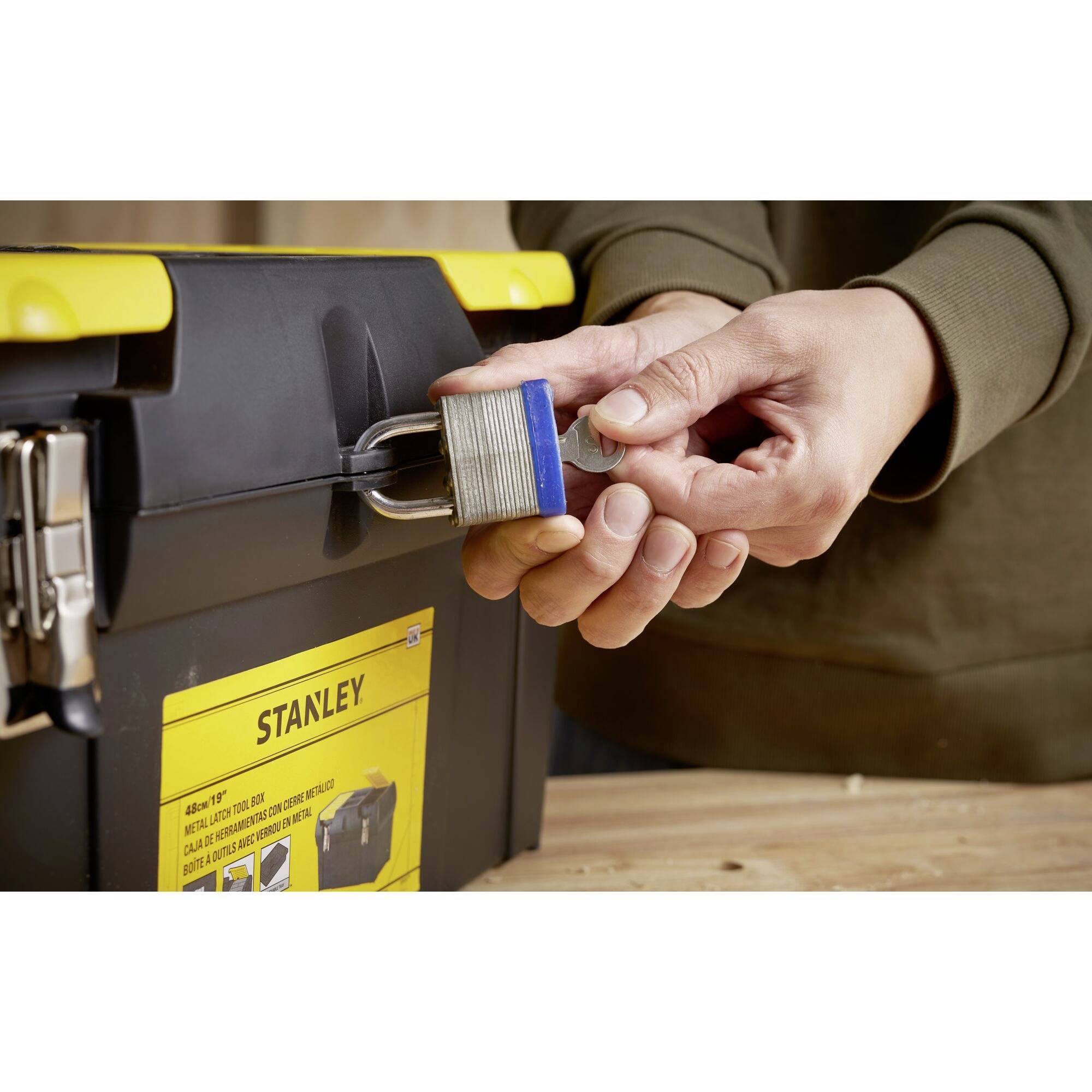 Une main ferme une boîte à outils noire avec un cadenas. La boîte comporte des détails jaunes et une étiquette.