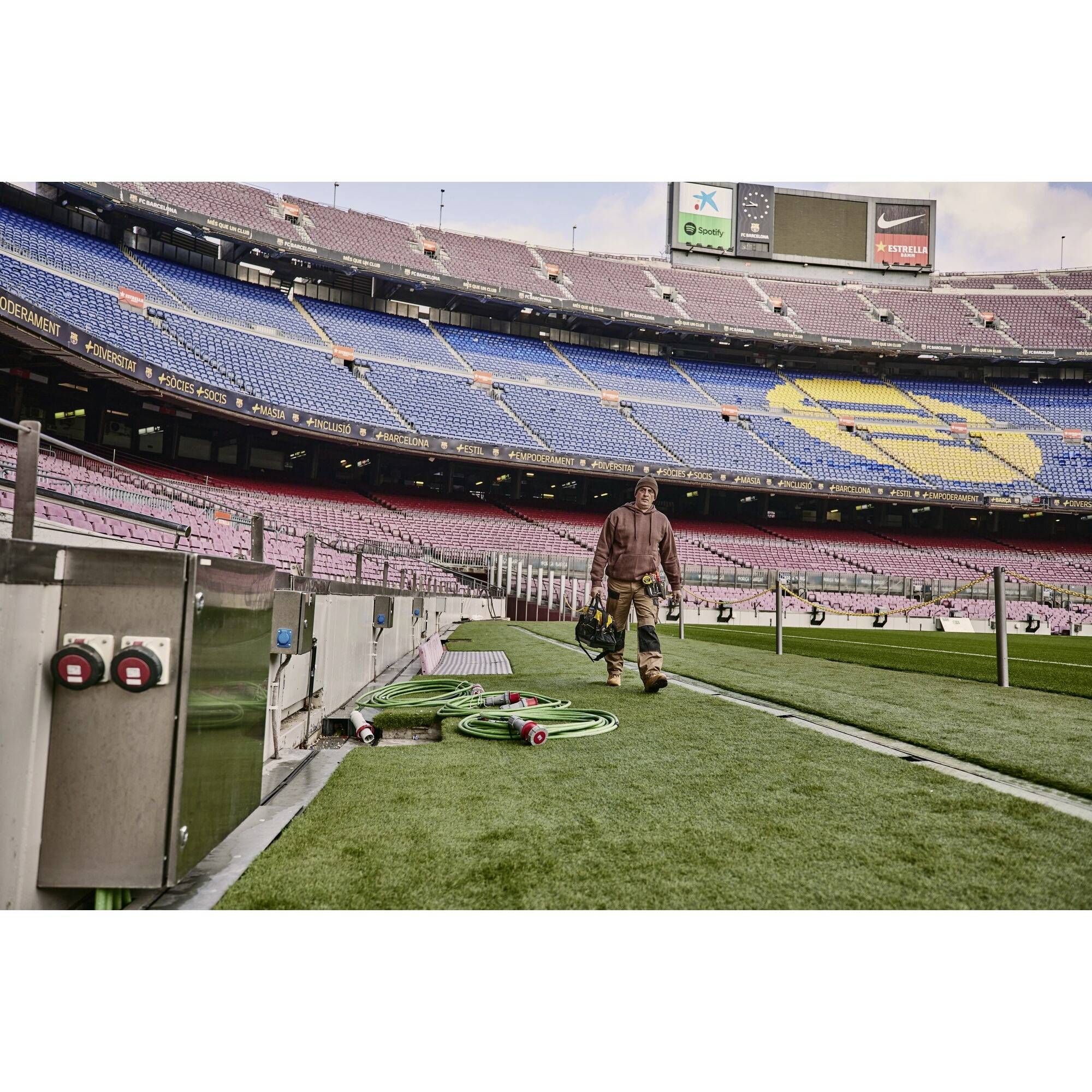 Un stade avec des rangées de sièges vides en bleu et rouge. Un ouvrier transporte de l'équipement sur un gazon vert, plusieurs tuyaux sont posés au sol.