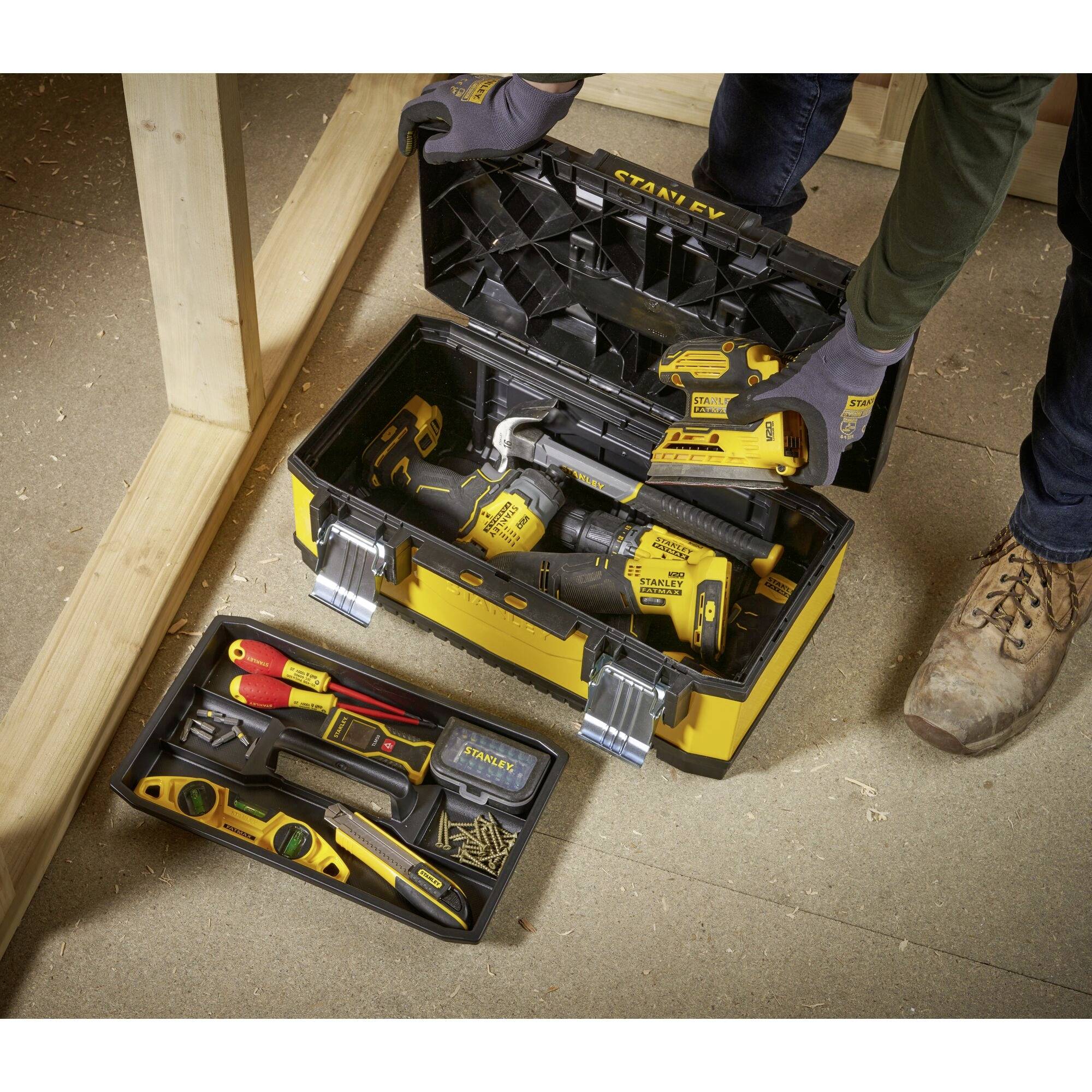 Une personne ouvre une boîte à outils jaune sur un plancher en bois. À l'intérieur se trouvent une visseuse, des tournevis et d'autres outils.