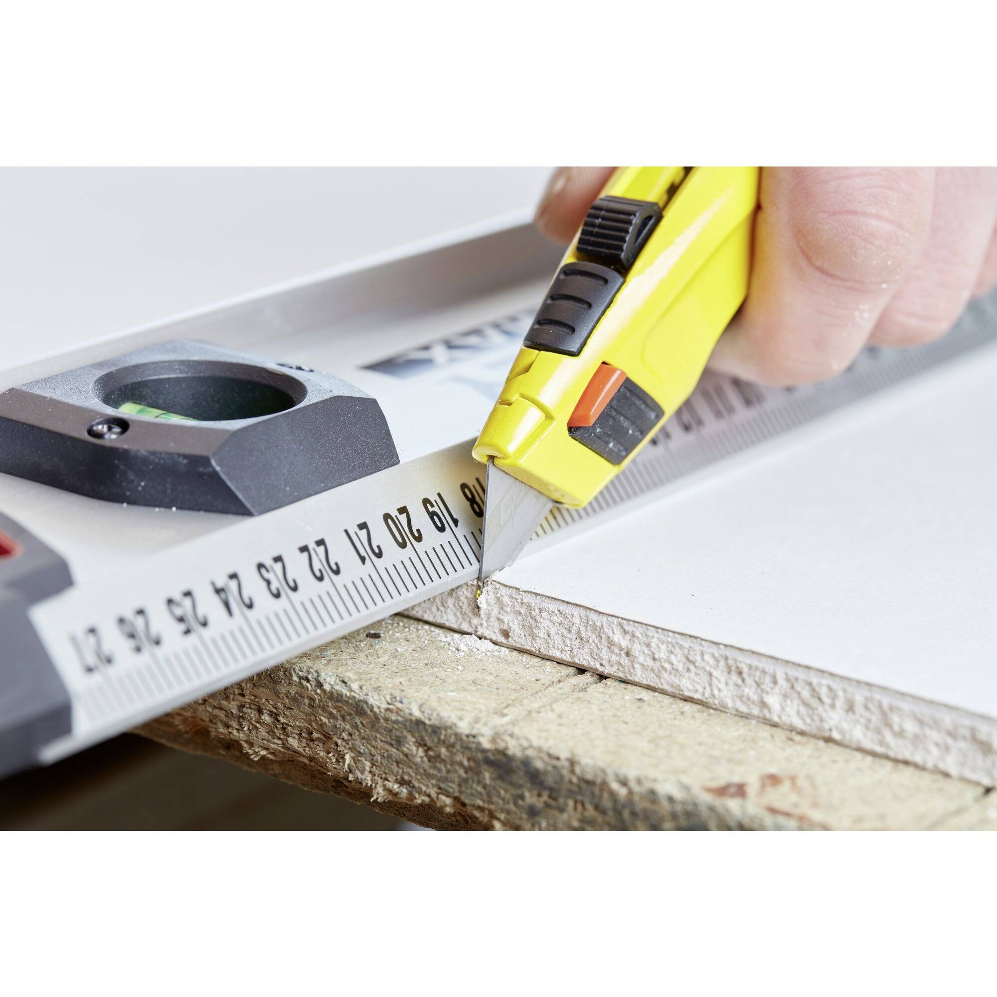 Une main coupe une épaisse plaque avec un cutter jaune le long d'une règle. La règle indique des mesures en centimètres.