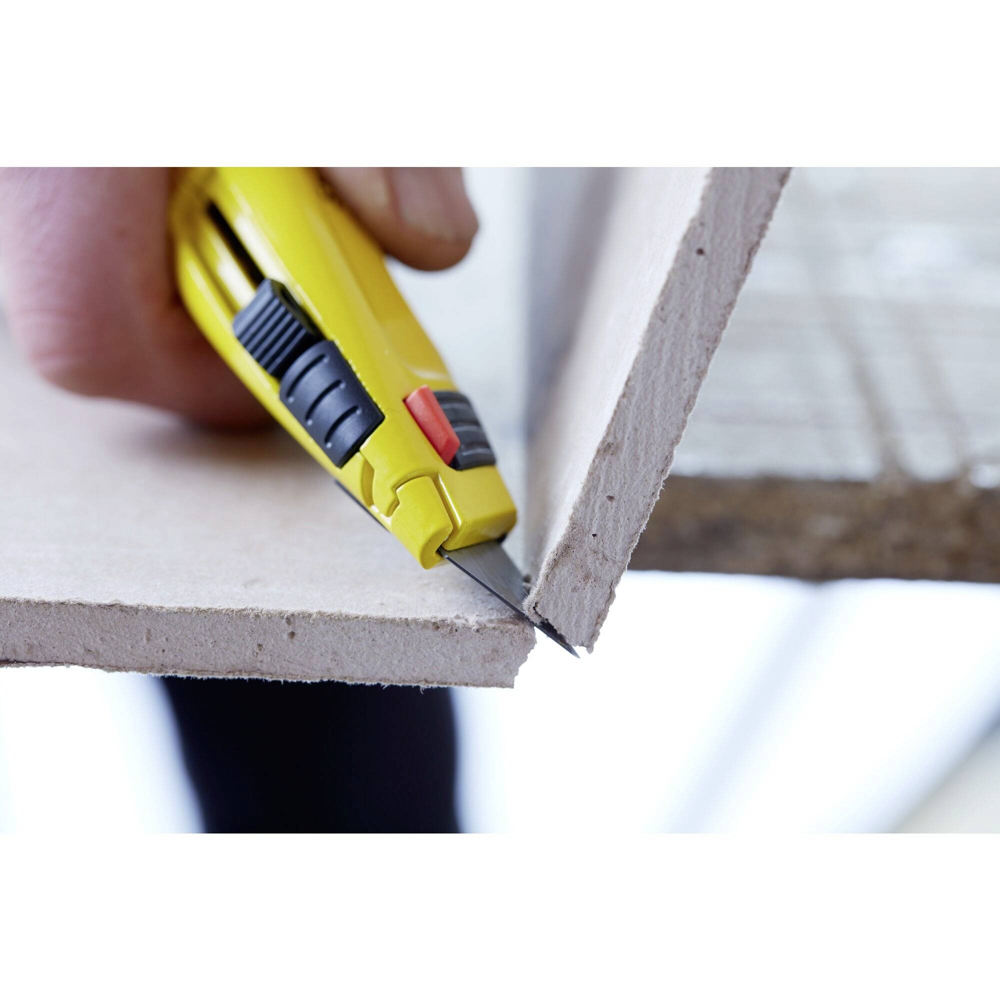 Une main coupe un morceau de contreplaqué le long du bord avec un cutter jaune.
