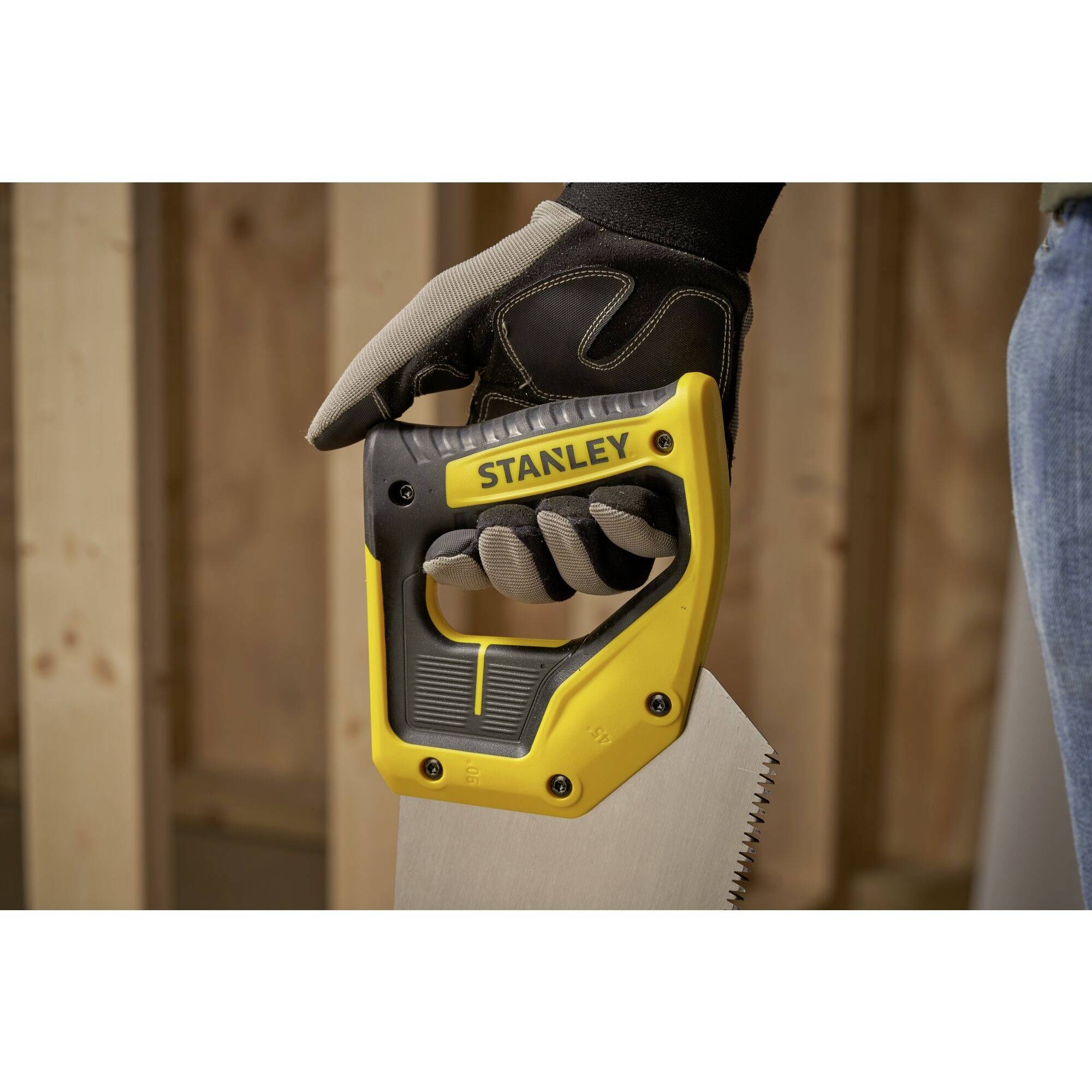 Une main tient une scie Stanley jaune, portée avec un gant de travail. Des poutres en bois sont visibles en arrière-plan.