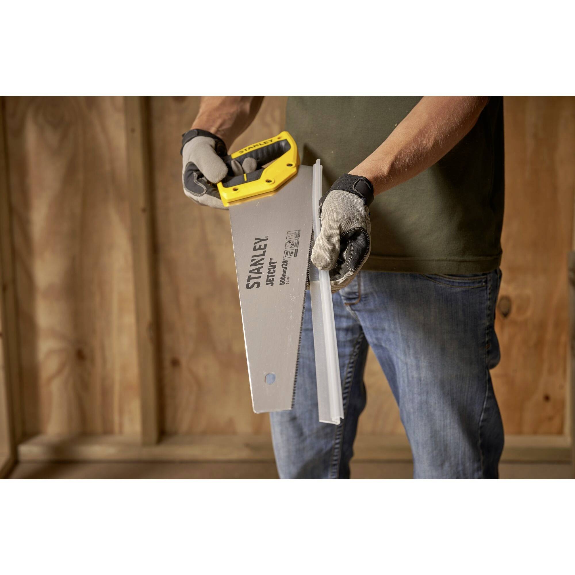 Un homme tient une scie à main et coupe un morceau de bois. La scie a une poignée jaune. L'arrière-plan montre un mur en bois.