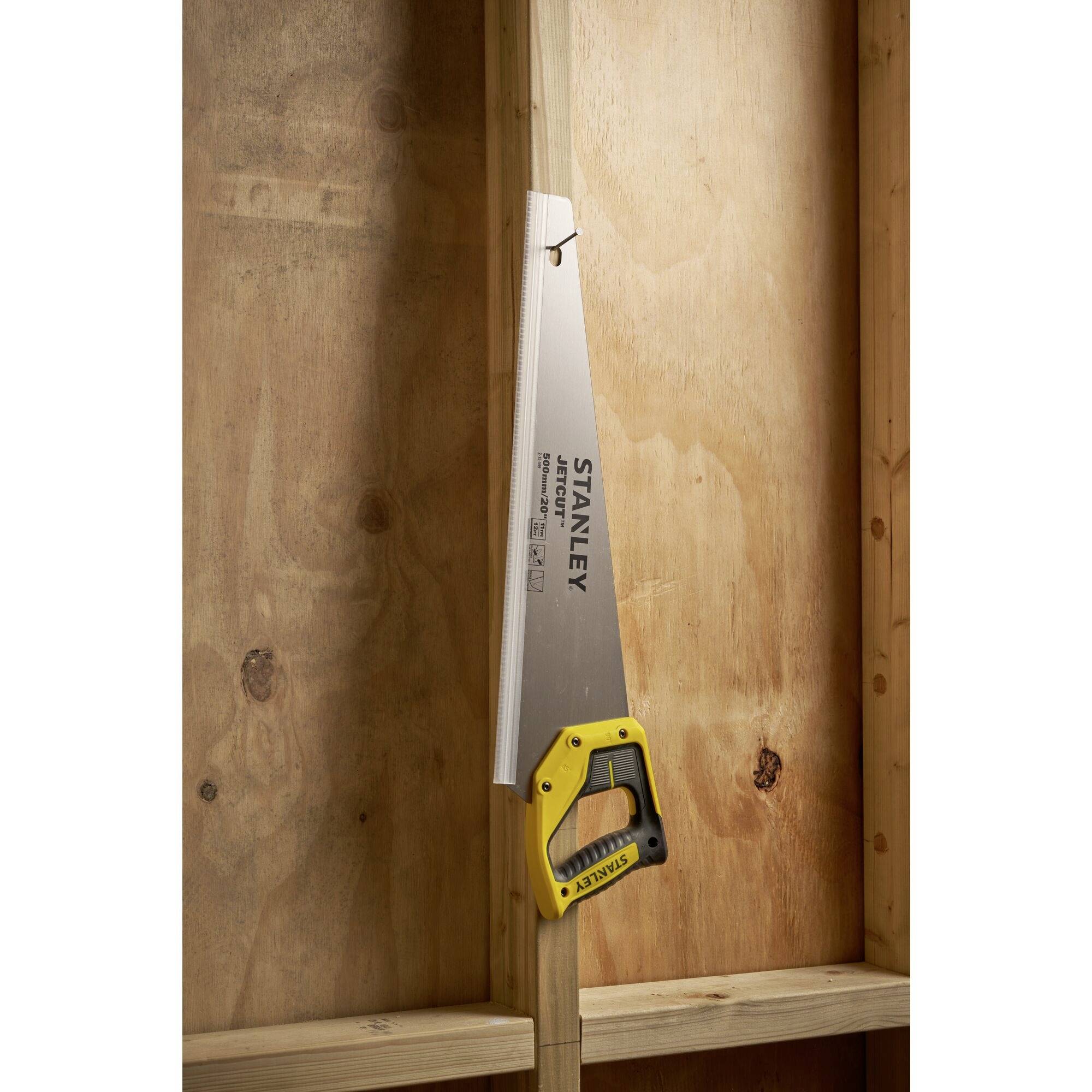 Une scie à main avec une poignée jaune et une lame argentée est suspendue à un mur en bois, illustrant le rangement des outils.