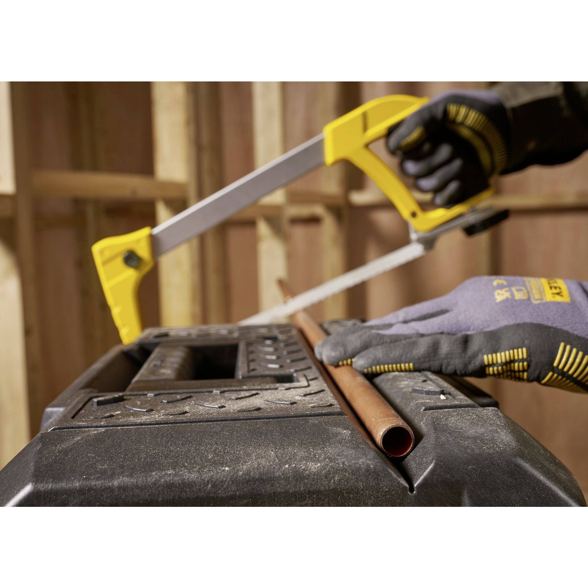 Une personne portant des gants de travail scie un tuyau de cuivre avec une scie métallique jaune, posé sur une table de travail noire.