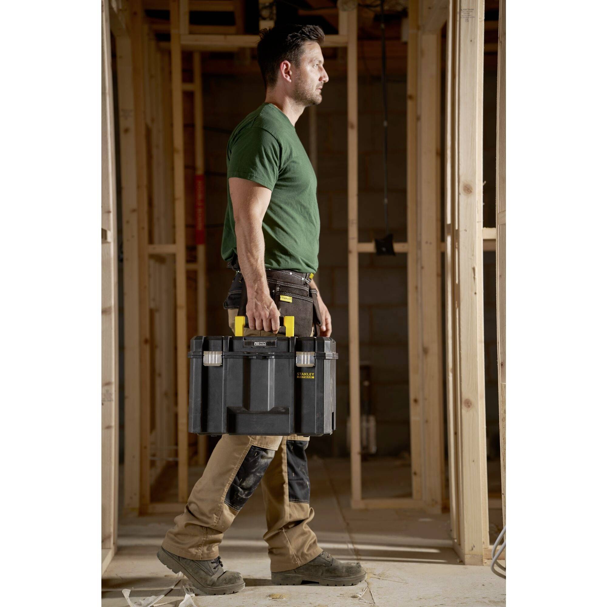 Un homme en tenue de travail transporte une boîte à outils à travers une pièce inachevée avec des murs à ossature bois.