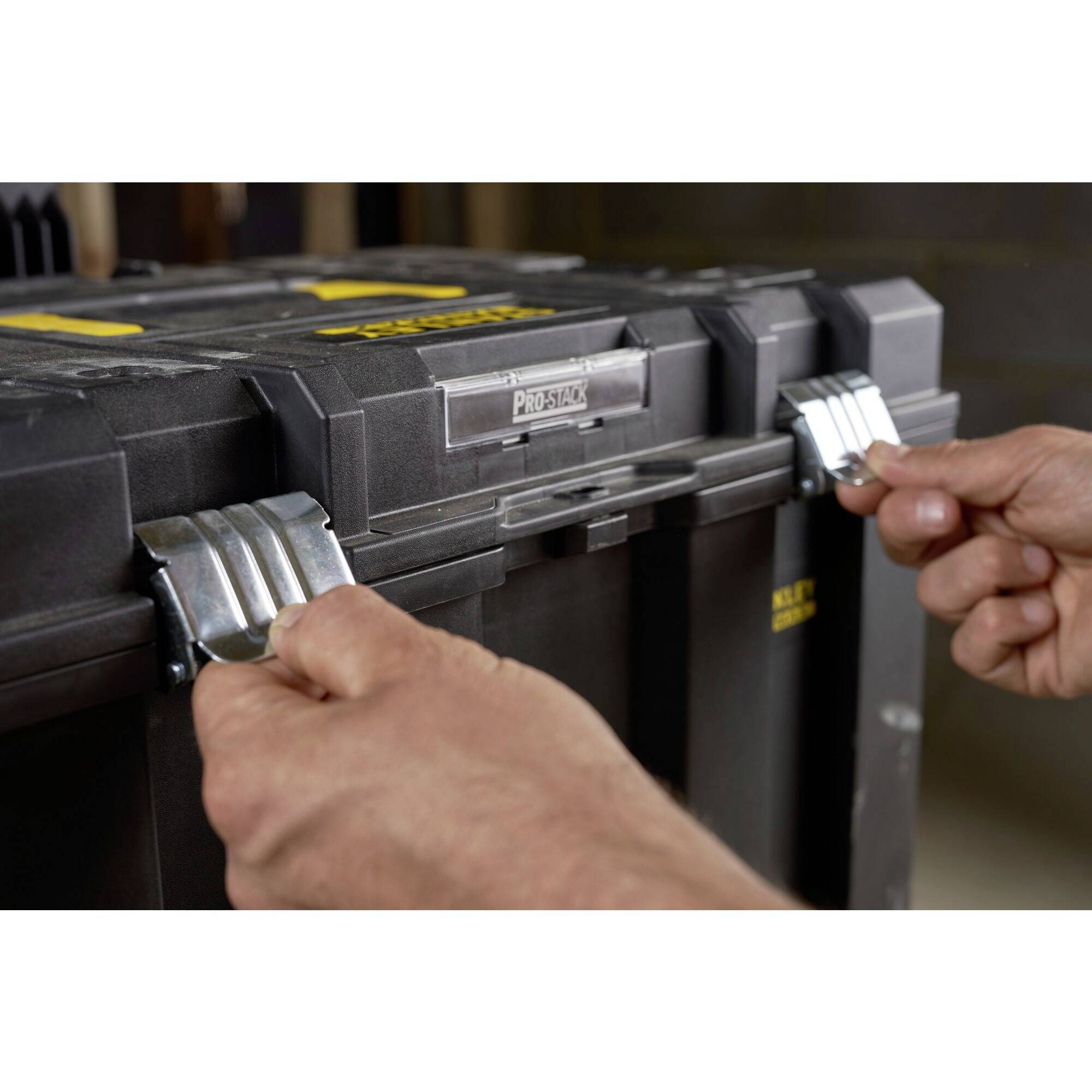 Des mains ouvrent les fermoirs métalliques d'une grande boîte à outils noire. La boîte présente des touches jaunes et argentées.