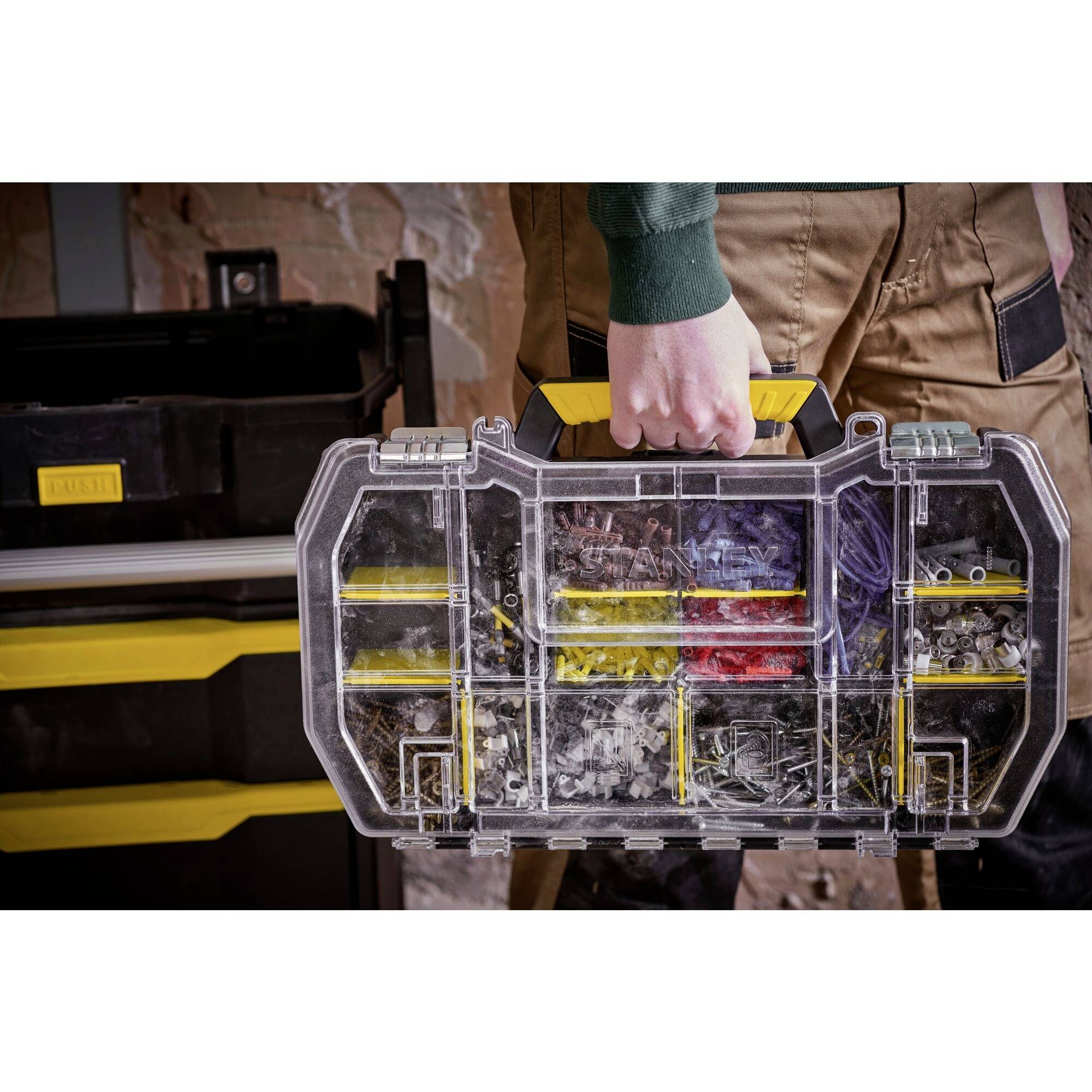 Une personne tient une boîte à outils transparente avec une poignée jaune, remplie de vis et de petites pièces. À l'arrière-plan, se trouve une caisse à outils.