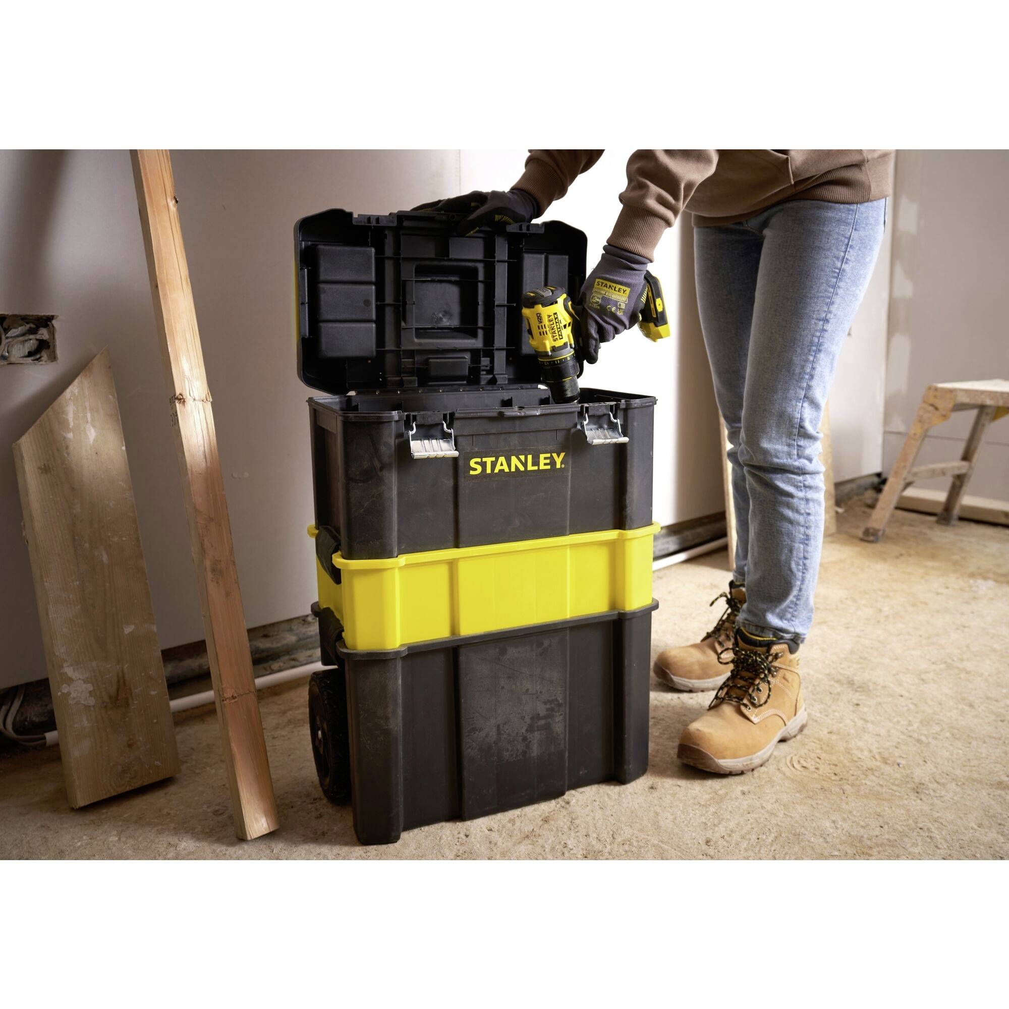 Une personne se tient dans une pièce, tenant une perceuse et rangeant des outils dans une grande boîte à outils Stanley jaune et noire.