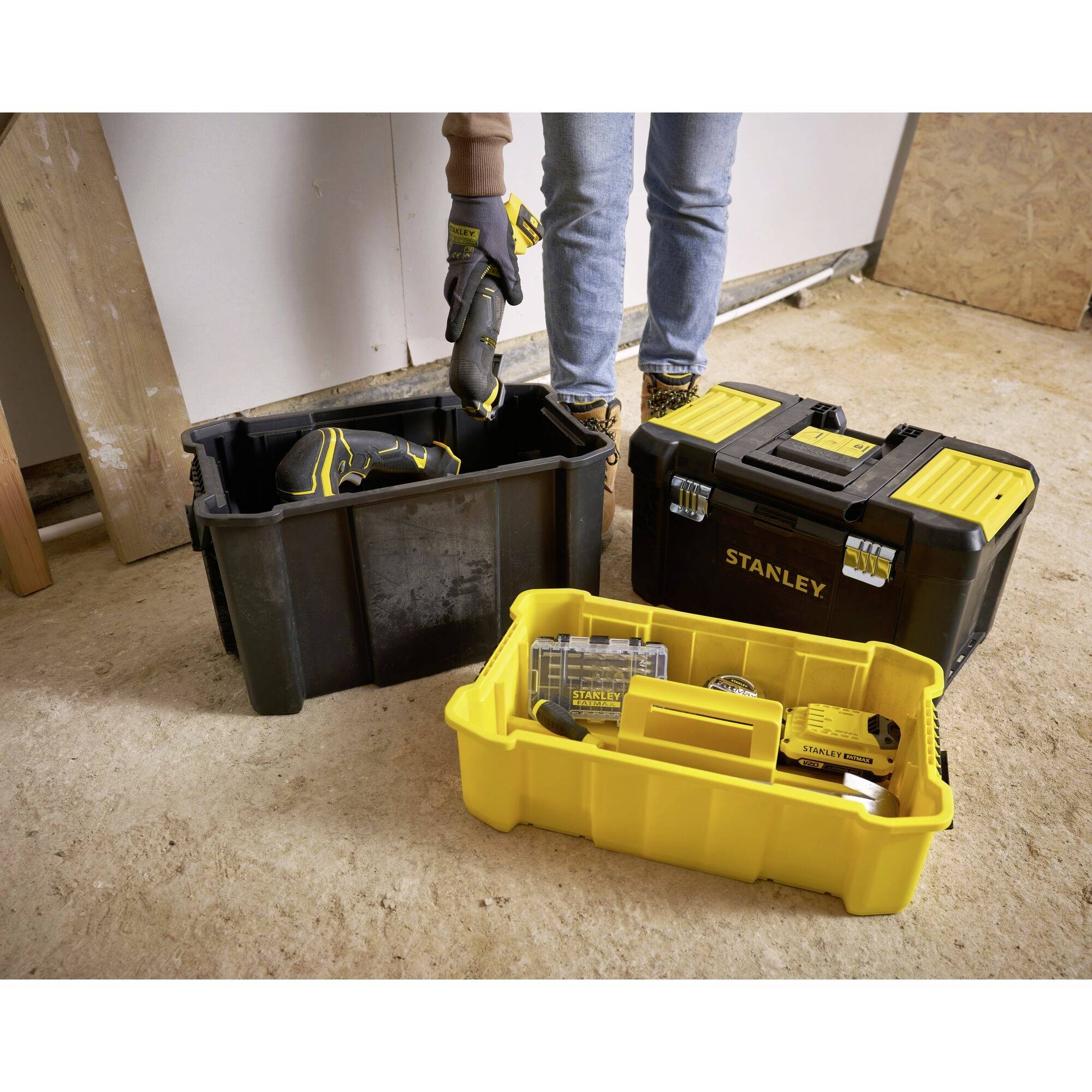 Une personne sort des outils d'une boîte à outils jaune et noire posée sur le sol, un visseuse-perceuse sans fil est clairement visible.