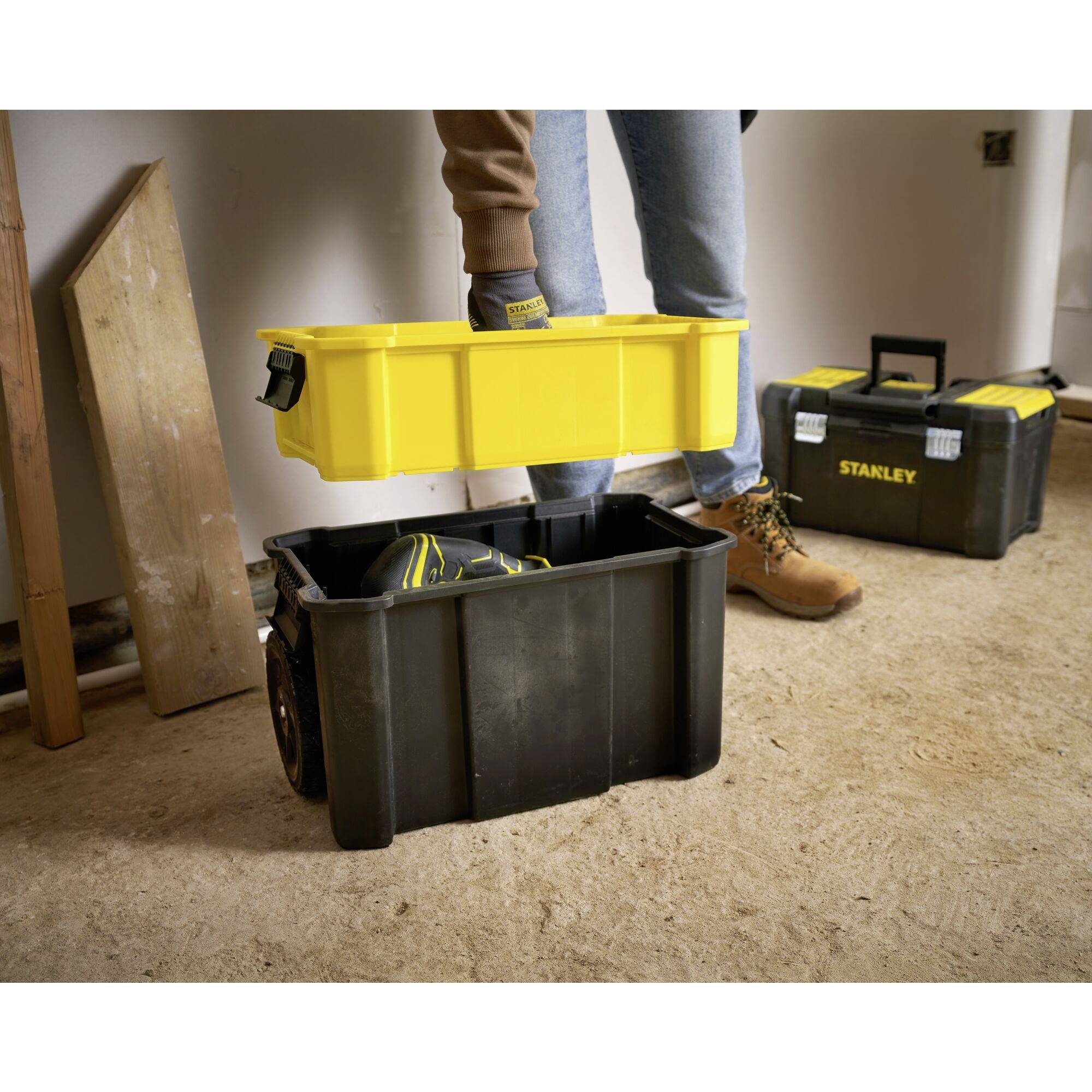 Une personne porte une boîte à outils jaune dans une pièce avec des planches de bois au sol. À l'arrière-plan, une autre boîte à outils noire est visible.