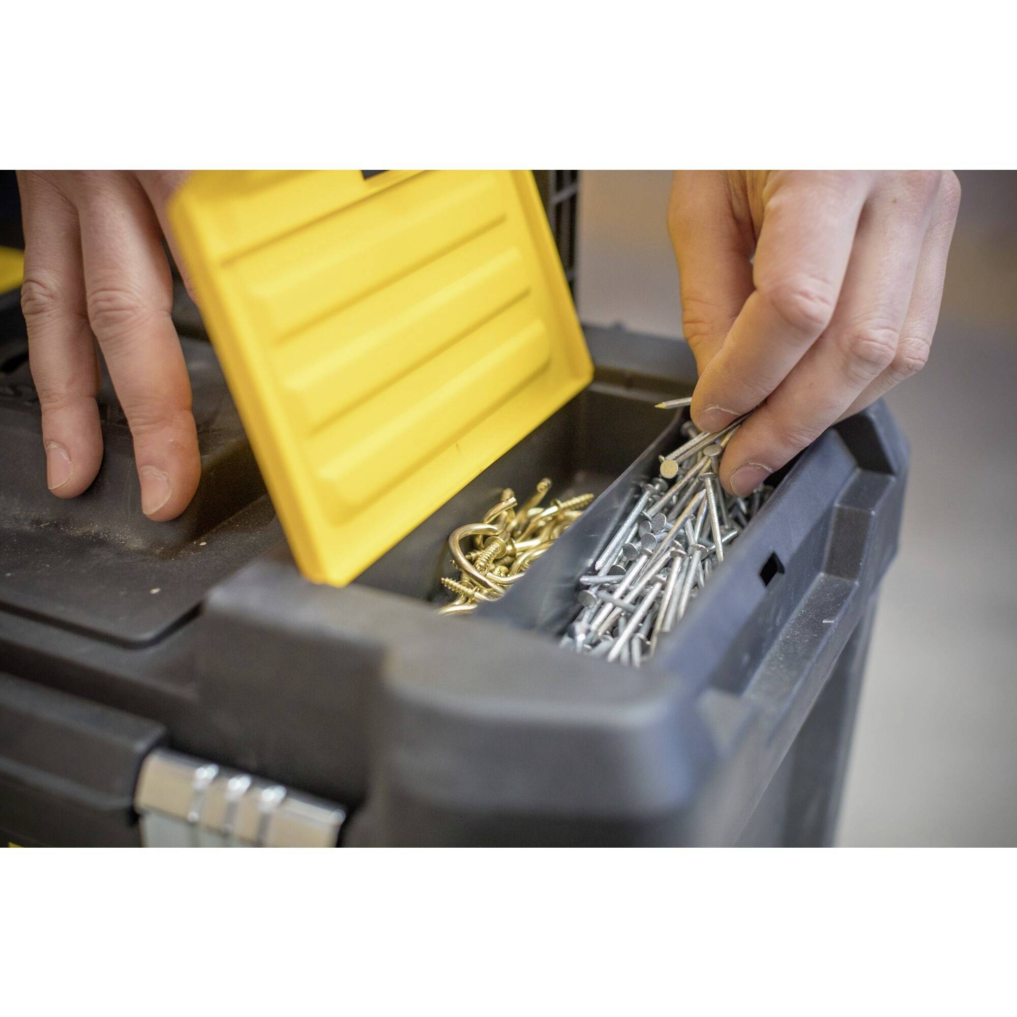 Deux mains saisissent des clous dans une boîte à outils jaune. La boîte est partiellement ouverte et révèle différentes pièces métalliques.