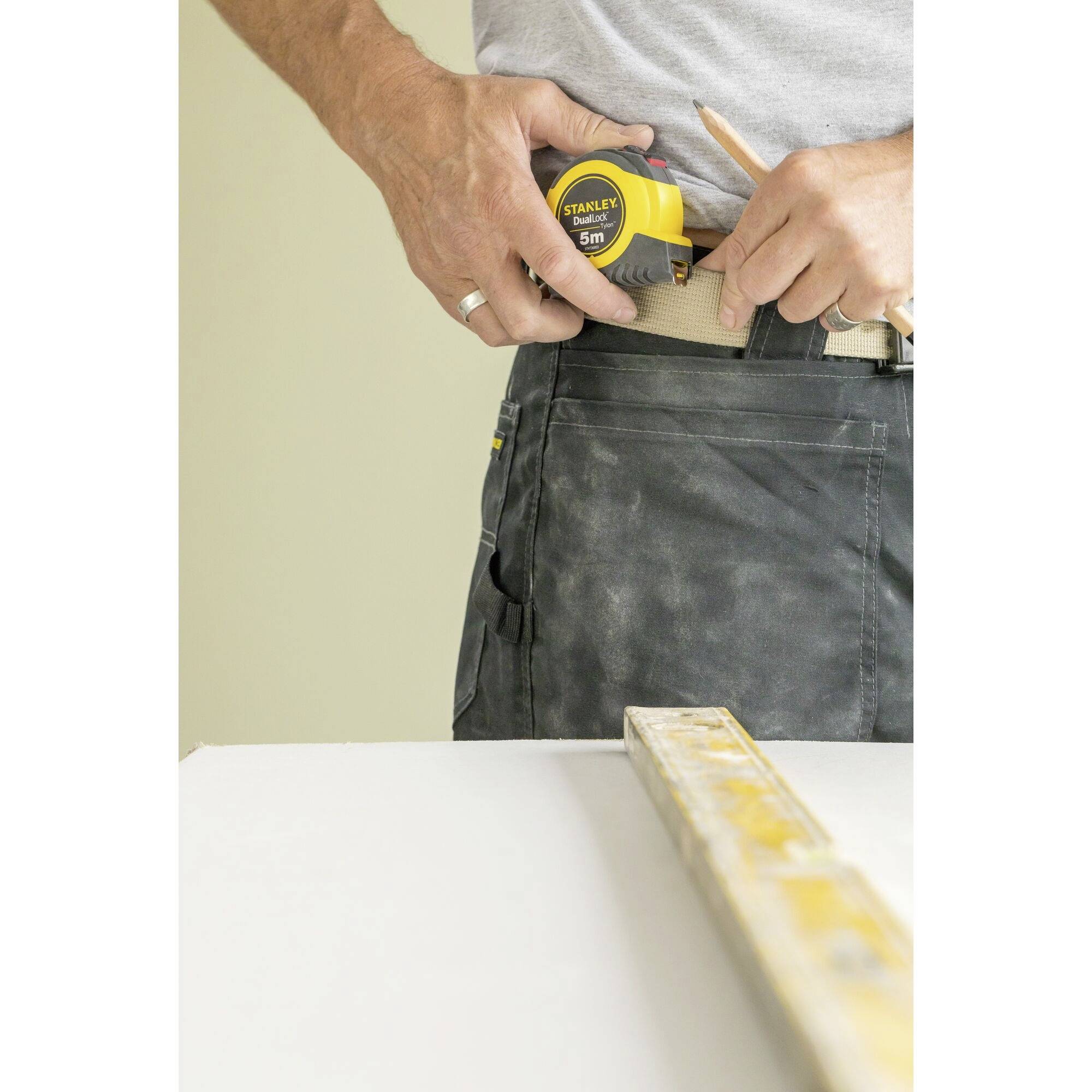 Une personne sort un mètre ruban d'un support fixé à sa ceinture, un mètre pliant est posé sur une table au premier plan.