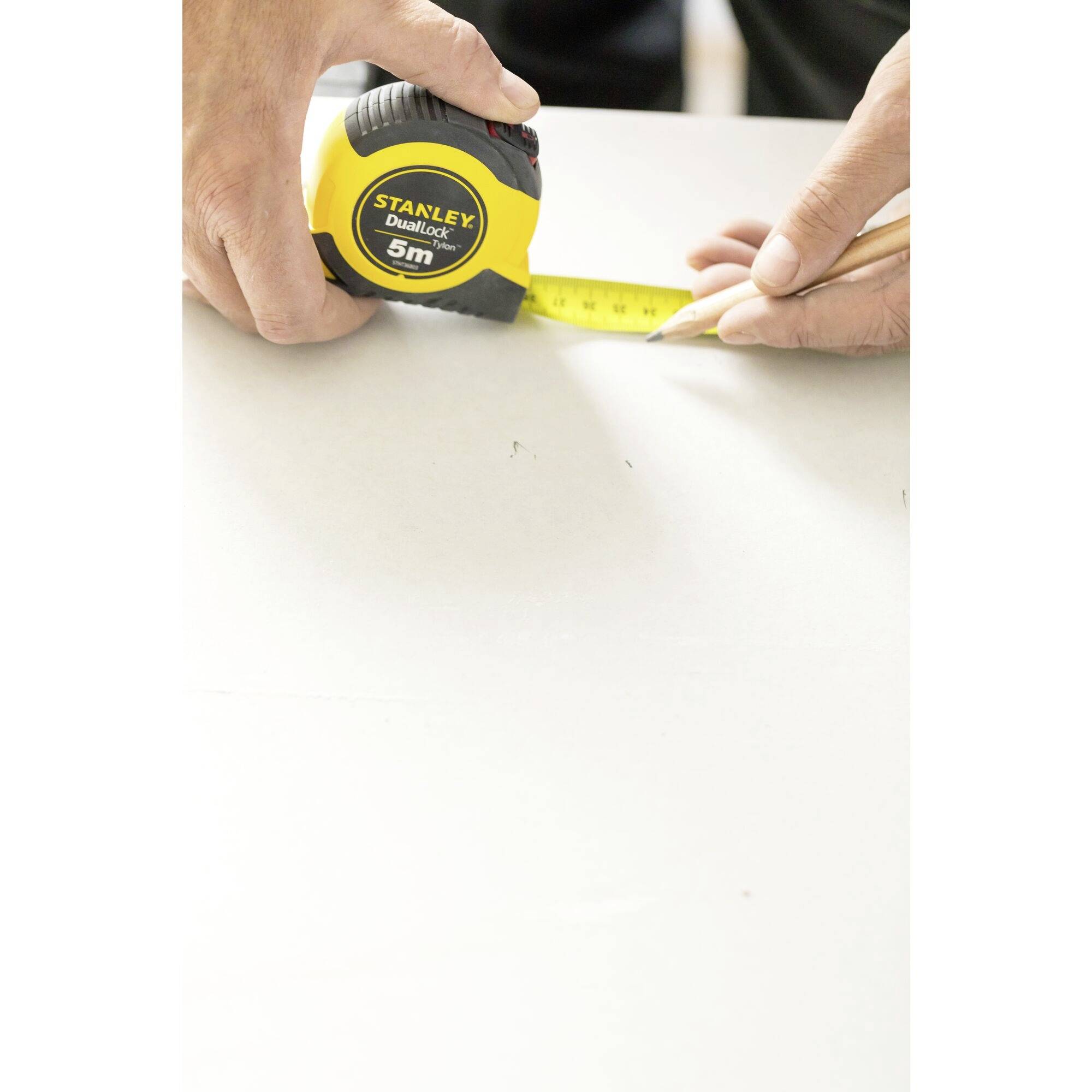 Une main tient un mètre ruban et mesure avec un crayon sur une surface blanche. Unité de mesure indiquée en mètres.