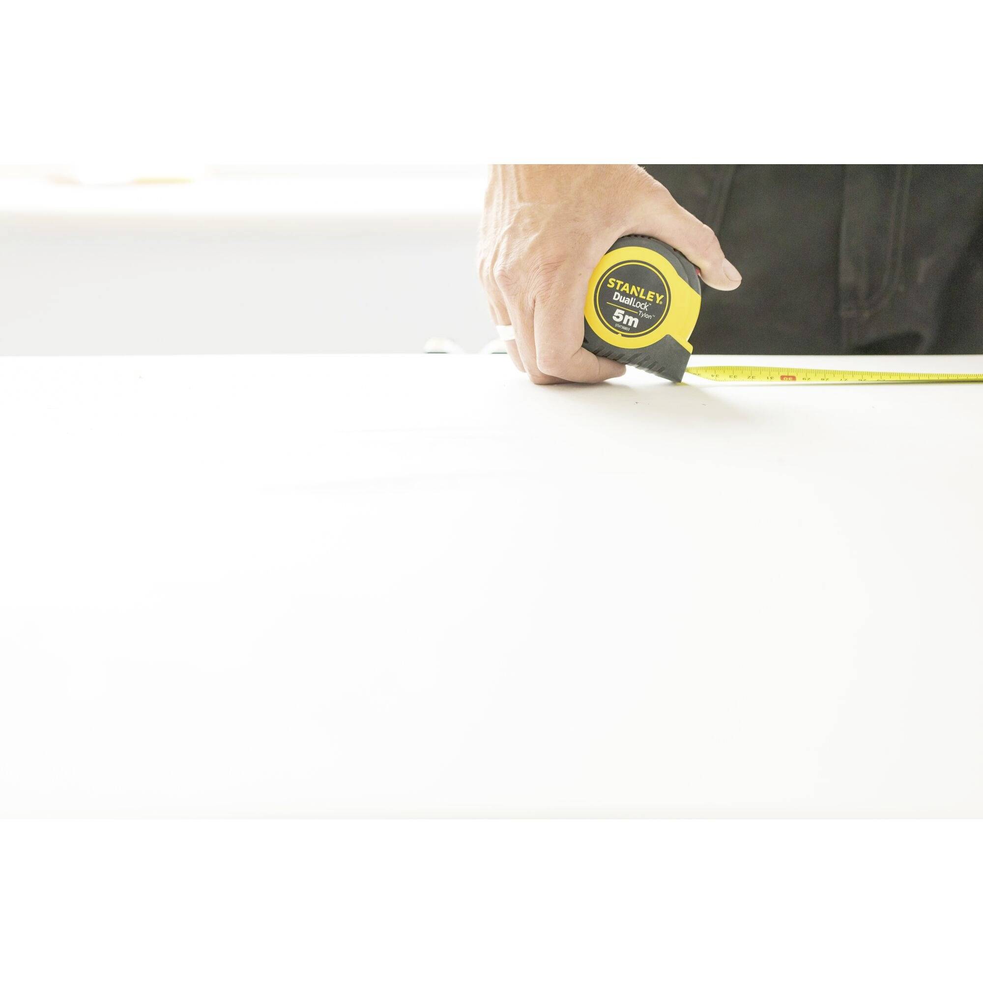 Une main tient un ruban à mesurer jaune et mesure une surface blanche. Le ruban indique 5 mètres.