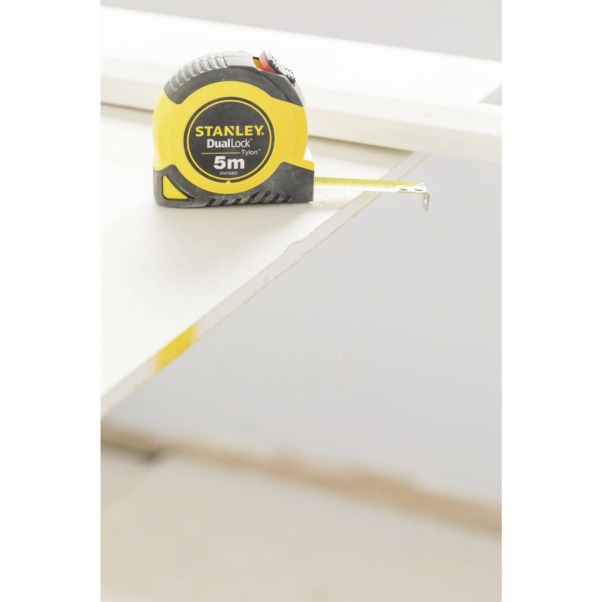 Un mètre ruban jaune et noir portant l'inscription 'Stanley DualLock 5m' est posé sur une planche blanche.