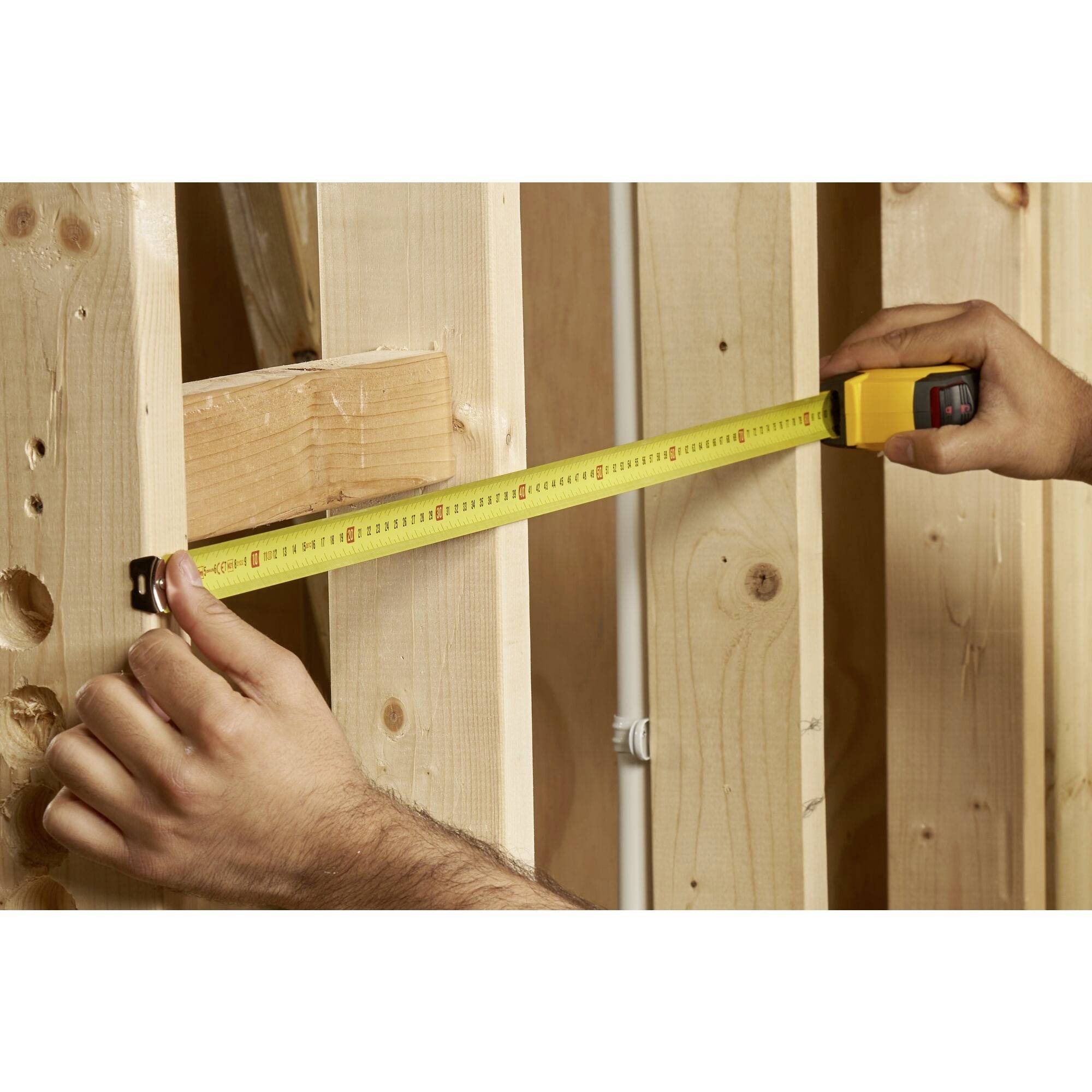 Une personne mesure la distance entre deux cadres en bois à l'aide d'un mètre ruban, probablement lors de travaux de construction ou de rénovation.