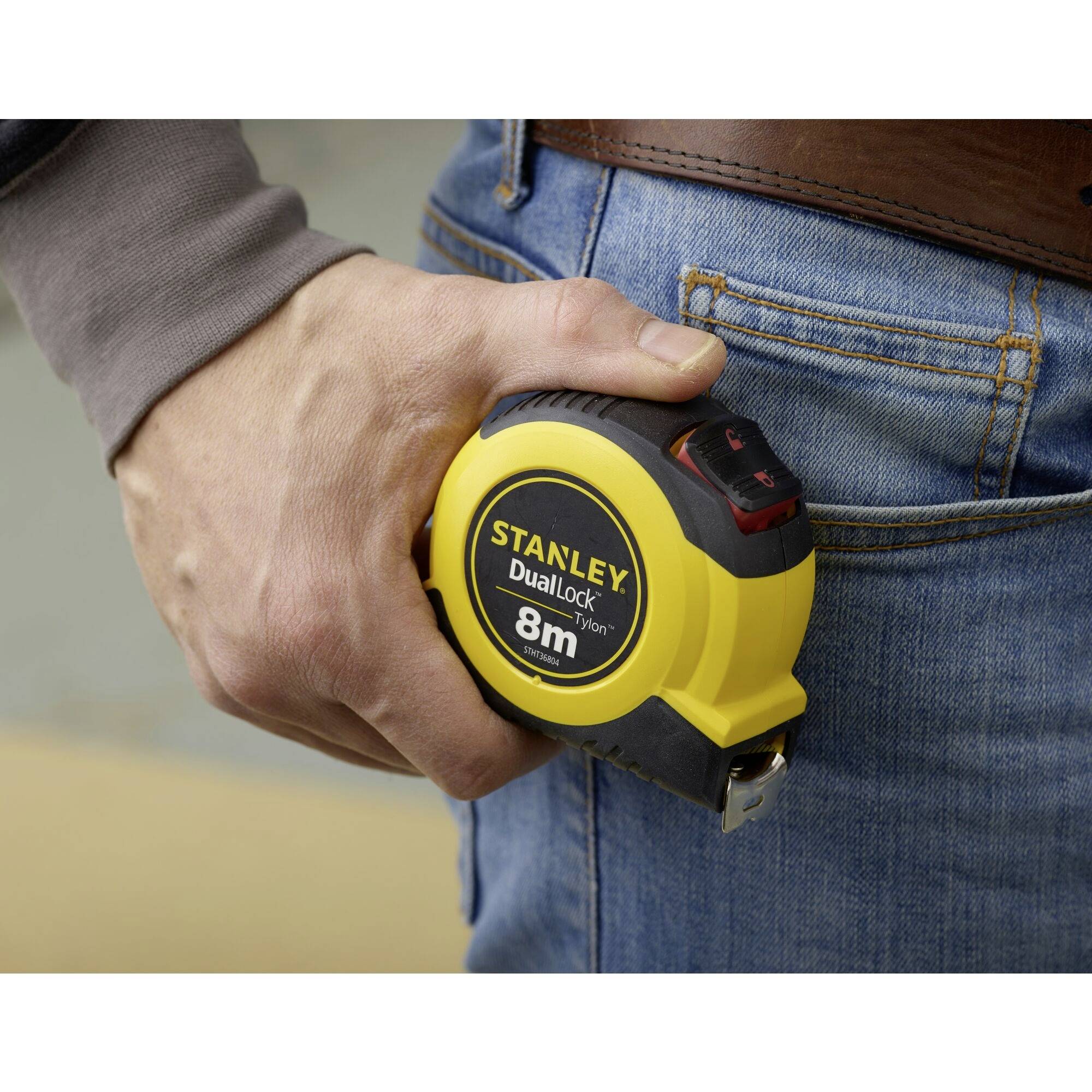 Une personne tient un mètre ruban jaune portant l'inscription 'Stanley DualLock 8m', fixé à sa ceinture.