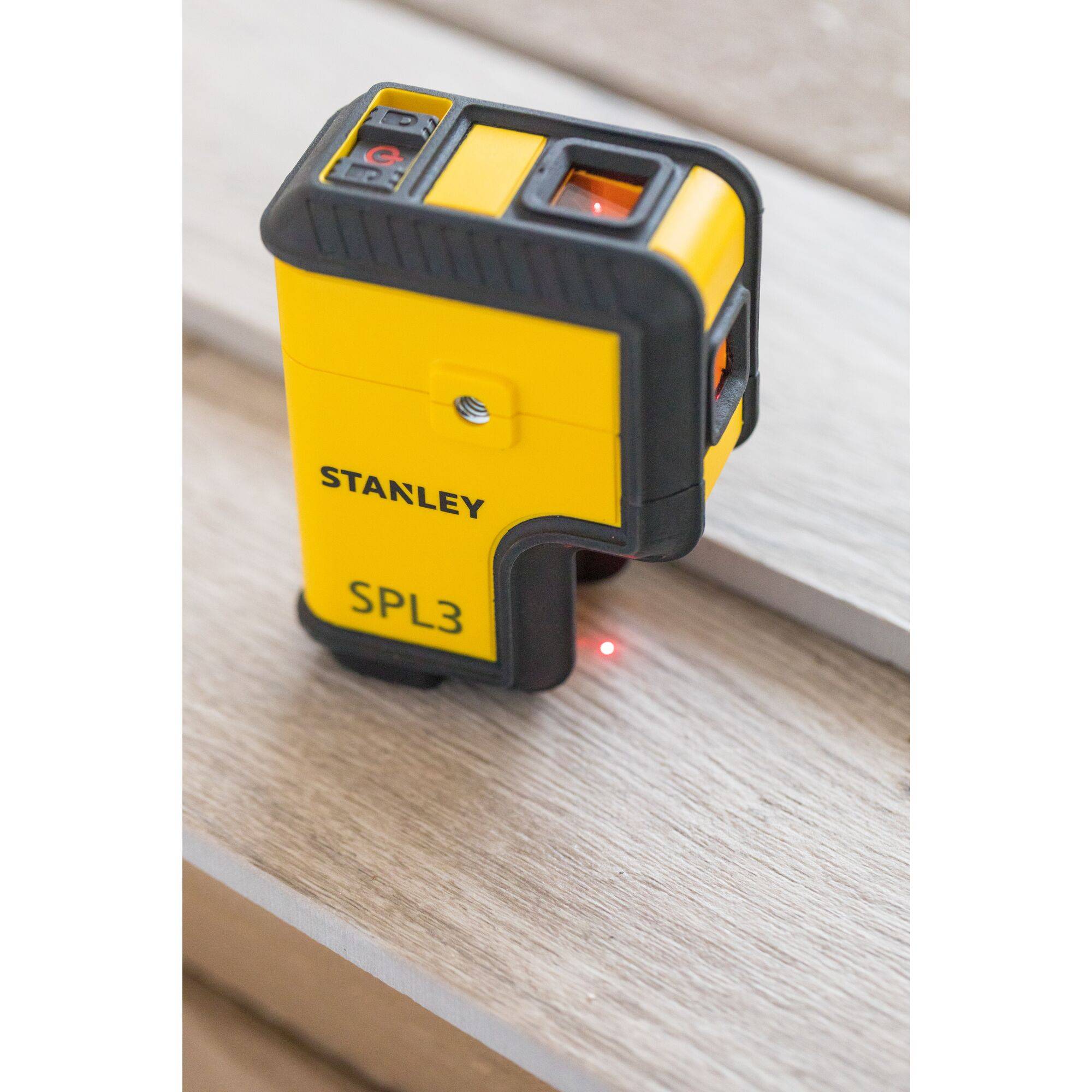 Un télémètre laser Stanley SPL3 jaune posé sur une surface en bois, affichant un point laser rouge.