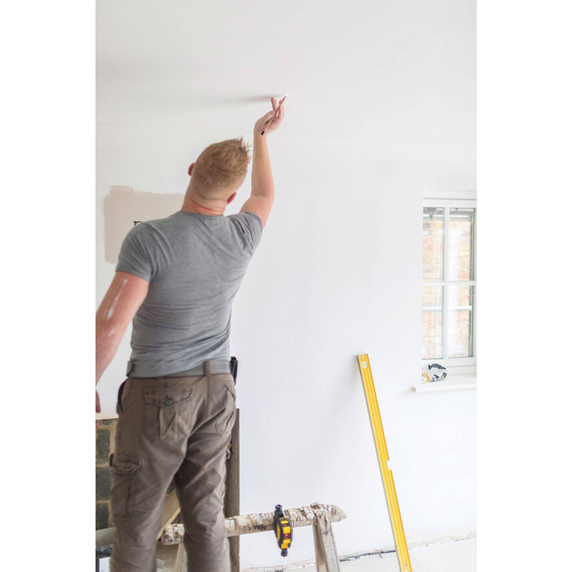 Un homme peint un mur en blanc. À côté de lui se trouve un niveau à bulle, ce qui suggère des travaux de rénovation ou de réaménagement.