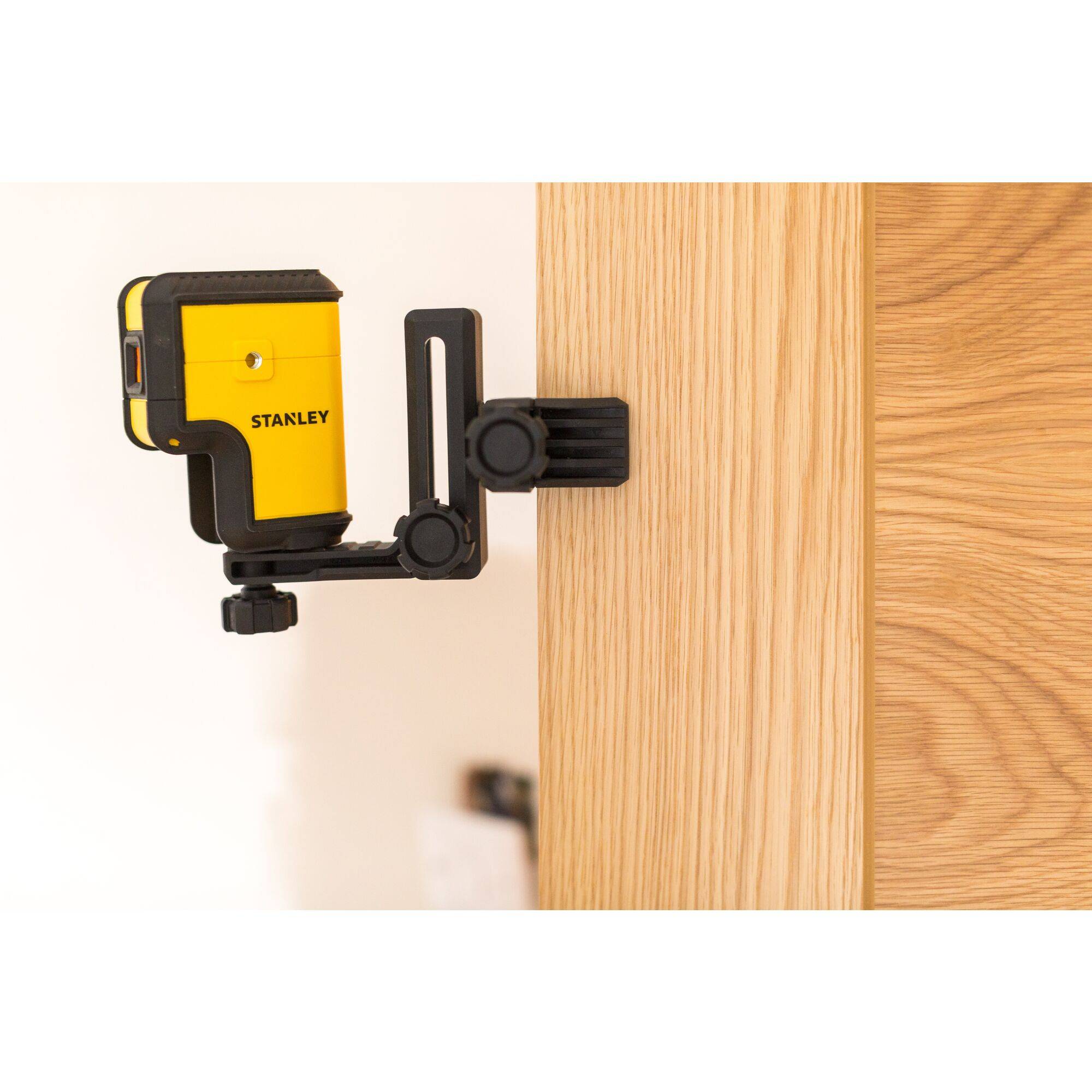 Mesureur de distance laser Stanley jaune, fixé avec un support sur un bord en bois. L'appareil est orienté vers la droite, avec un arrière-plan flou.