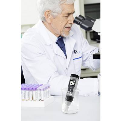 Un scientifique examine des échantillons sous un microscope dans un laboratoire. Au premier plan, un instrument de mesure est placé dans un bécher.