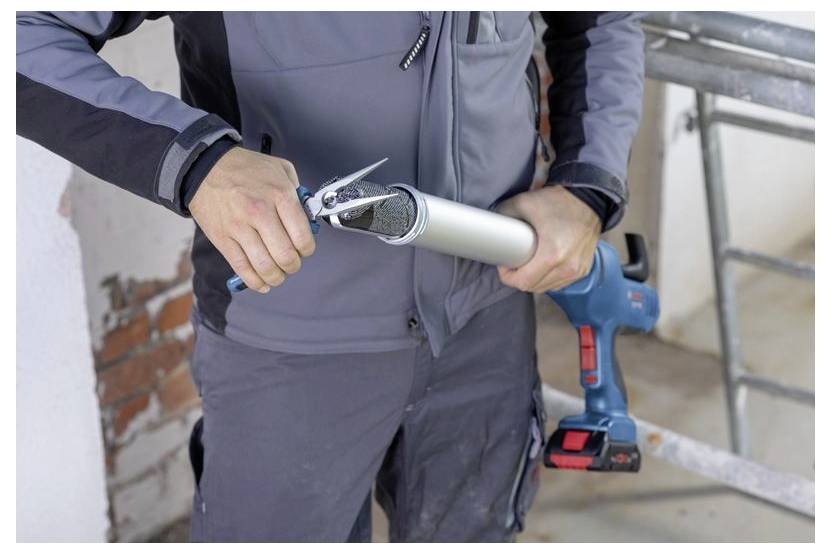 Une personne coupe l'embout d'un pistolet à calfeutrer avec un couteau utilitaire, en préparation de son utilisation. Elle se tient dans une pièce en cours de rénovation.