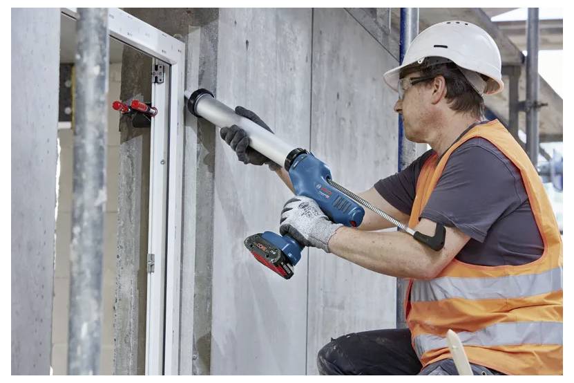 Un ouvrier du bâtiment portant un casque de chantier et un gilet orange utilise un pistolet à mastic pour sceller l'encadrement d'une fenêtre sur un chantier de construction.