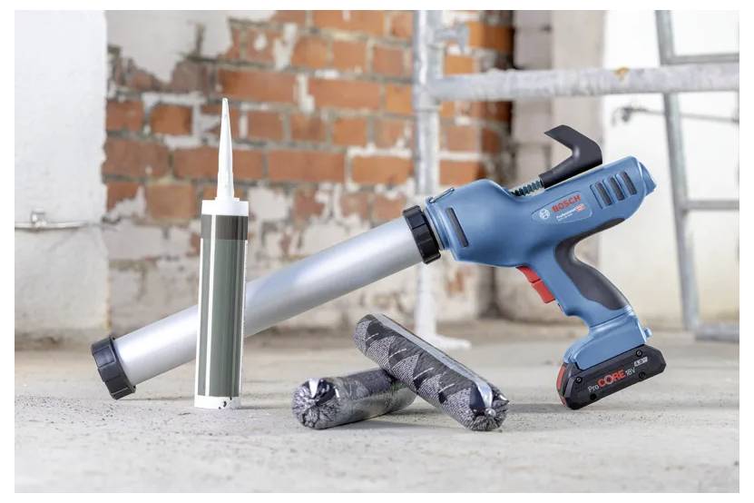 Un pistolet à calfeutrer sans fil avec sa batterie est posé sur un sol à côté de tubes et de saucisses de mastic. Le mur de briques apparent en arrière-plan suggère des travaux de rénovation.